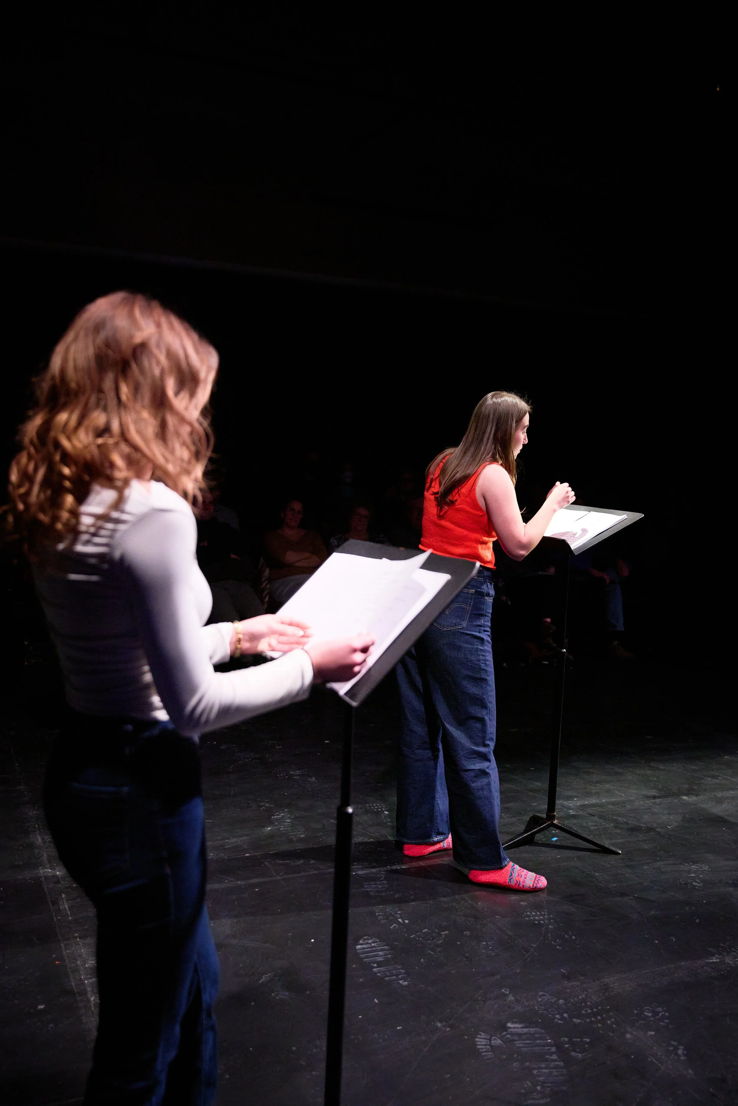 Deux femmes lisant des scripts ou des livres lors d'une performance ou d'une répétition sur scène, avec un public en arrière-plan dans l'obscurité.