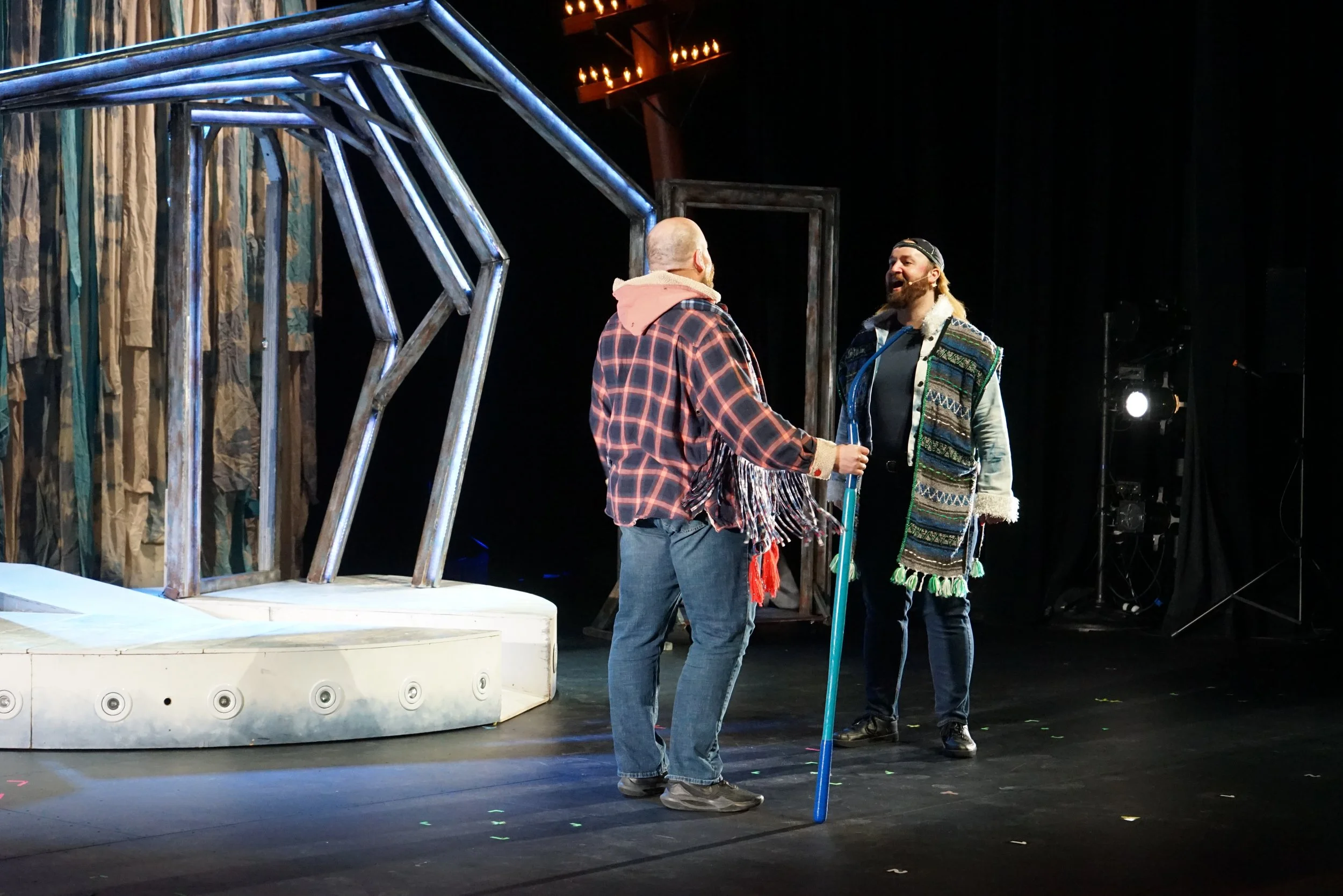 Deux hommes discutent sur scène, un tient une canne et porte un veston à motifs, l'autre un manteau à capuche avec une écharpe, dans un théâtre.