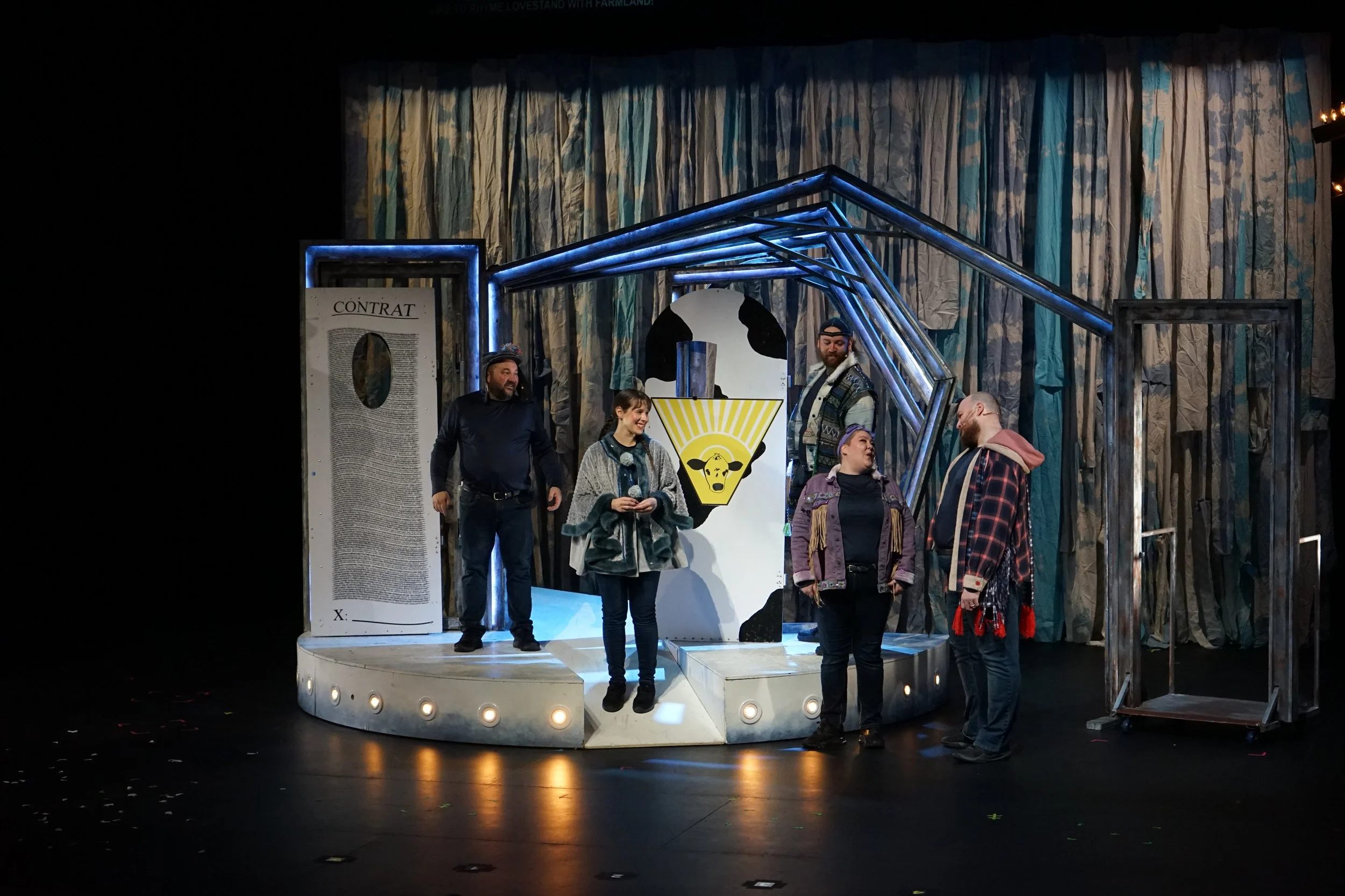 Six personnes sur une scène de théâtre, avec un décor abstrait en arrière-plan, discutant et souriant. La scène est éclairée, avec un panneau à gauche et un panneau central décoré d'une tête de vache et d'un logo jaune.