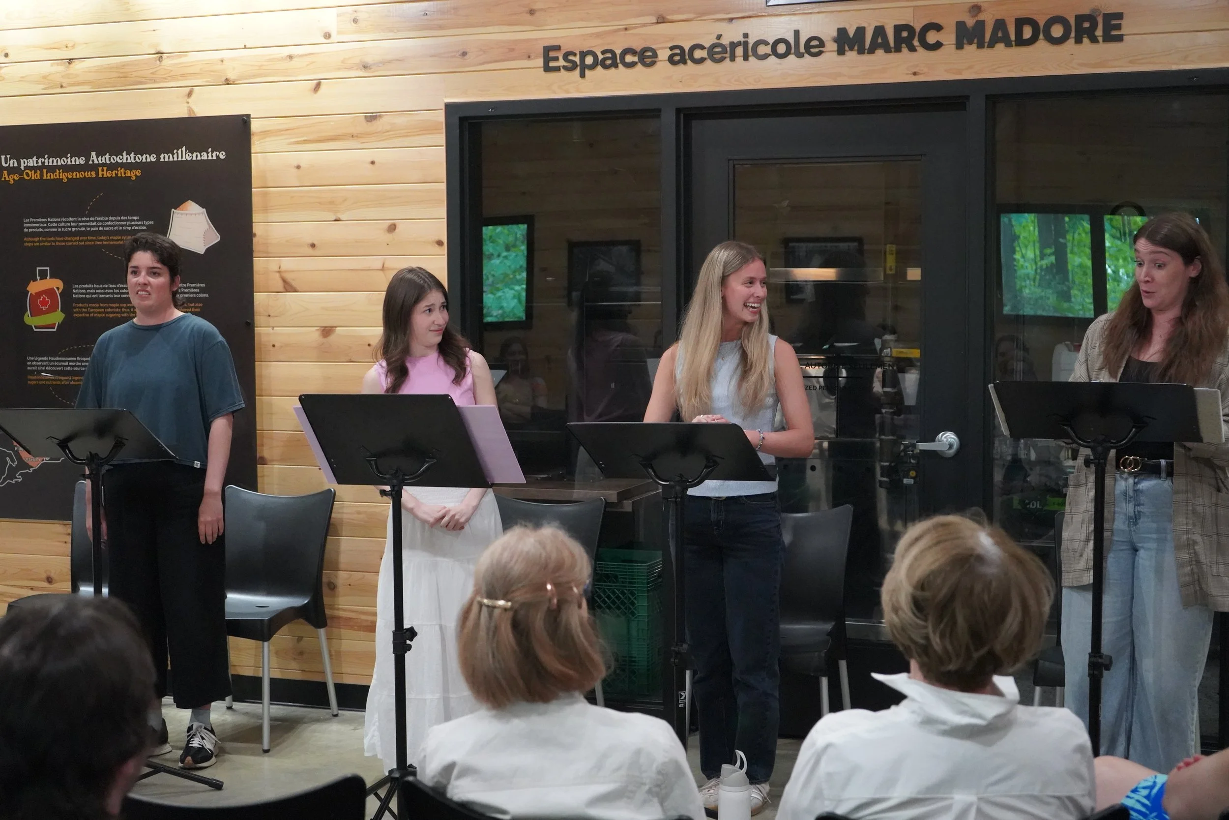 Groupe de quatre femmes et une fille parlant lors d'une présentation dans une salle avec murs en bois, devant un public.
