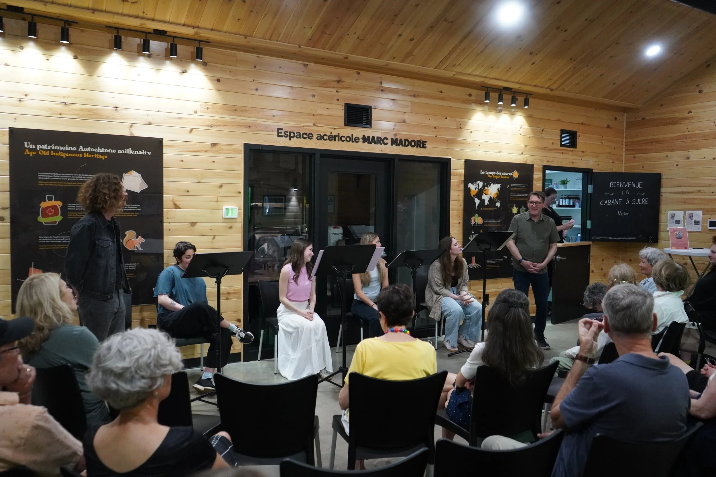 Une scène dans une salle en bois où plusieurs personnes assistent à une présentation ou une performance. Il y a un groupe de cinq jeunes assis devant un fond de personnes âgées, avec deux hommes debout. Des affiches informatives sont visibles sur le 