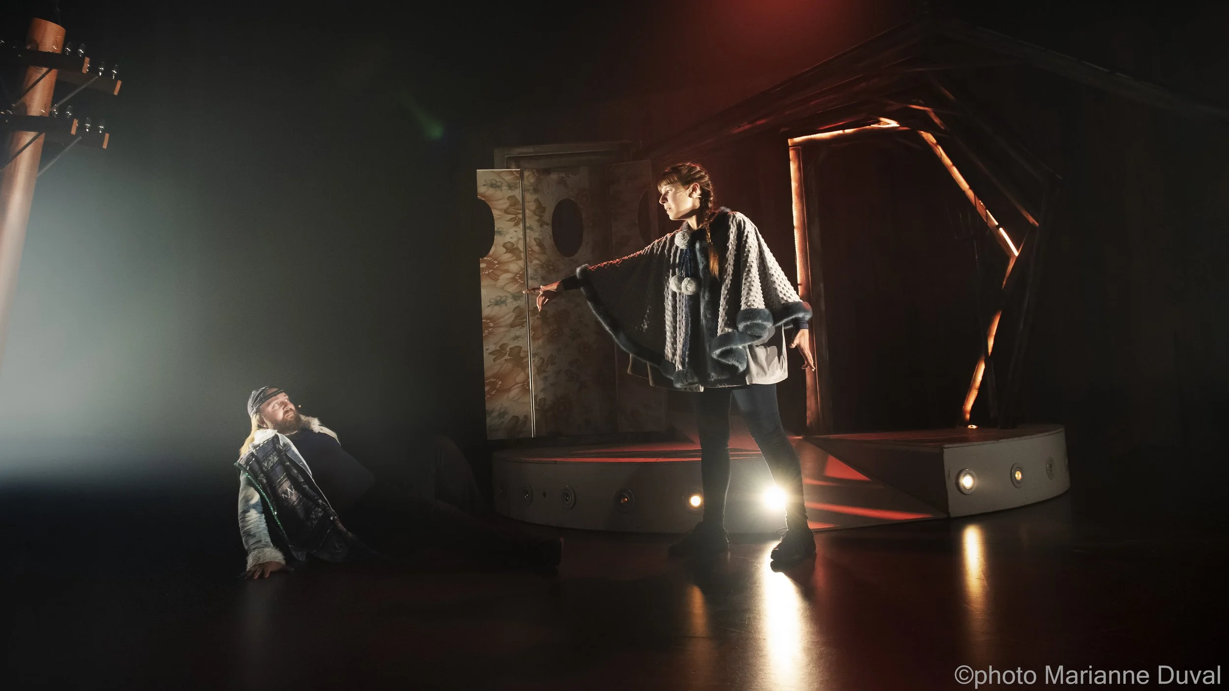 Une scène de théâtre avec deux acteurs, l'une debout avec un manteau à capuche et l'autre assis ou étendu sur le sol, dans une ambiance sombre avec une lumière dirigée sur eux.