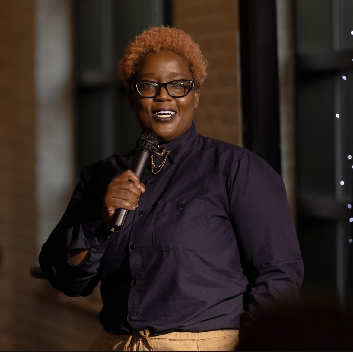 Femme noire avec cheveux courts et bouclés, portant des lunettes noires et une chemise noire, tenant un microphone, parlant lors d'un événement en intérieur.