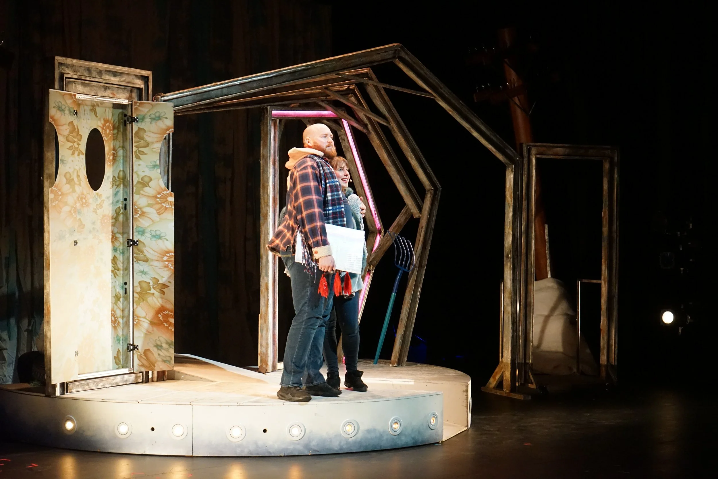 Trois personnes se tiennent sur un petit théâtre avec décor en bois, une porte, une pancarte de ferme, et des éléments de scène. L'une d'elles tient un document, elles semblent en pleine représentation ou conversation.