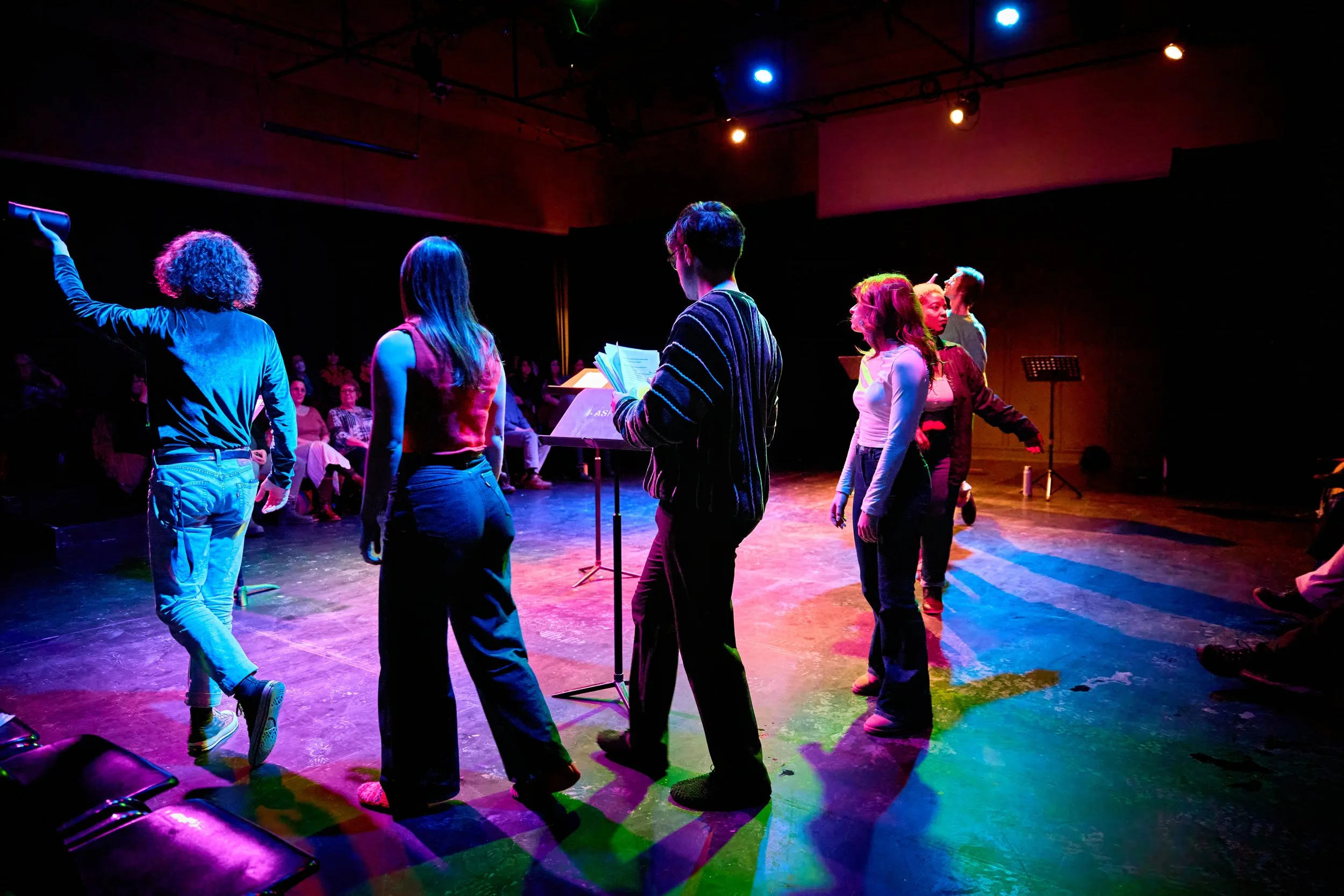 Groupe de personnes sur scène lors d'une représentation, avec un public assis en arrière-plan, sous un éclairage coloré.