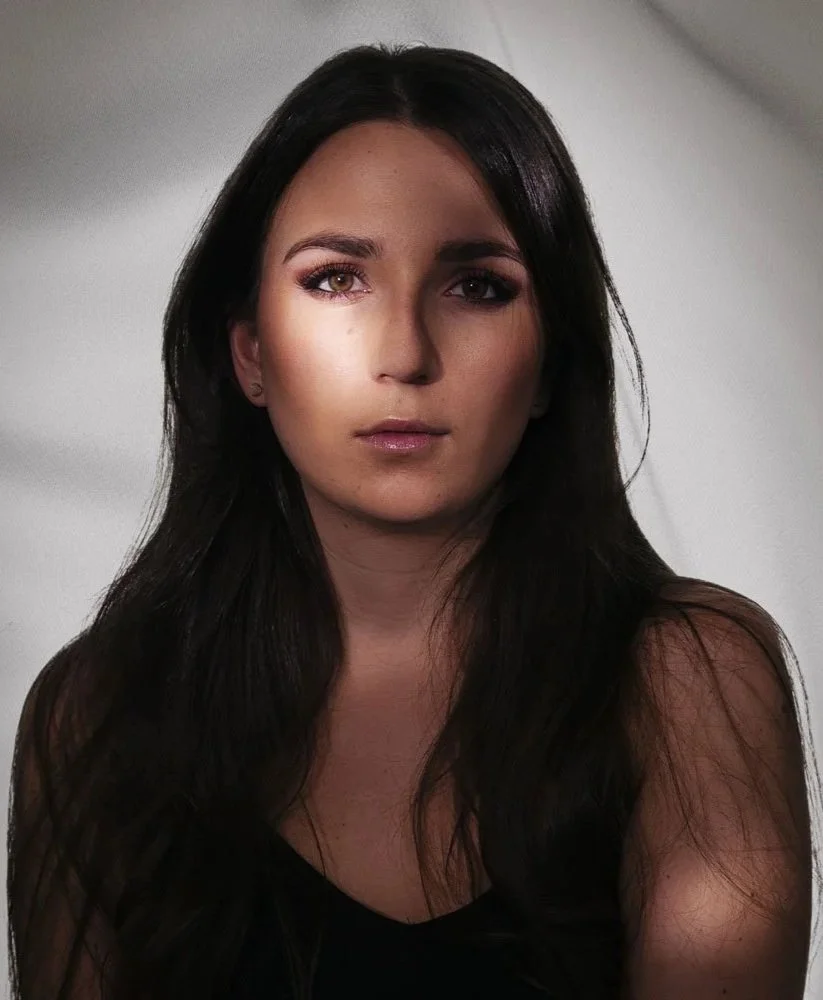 Une femme avec de longs cheveux noirs, maquillée, portant un haut noir, regardant vers l'objectif avec un fond neutre.