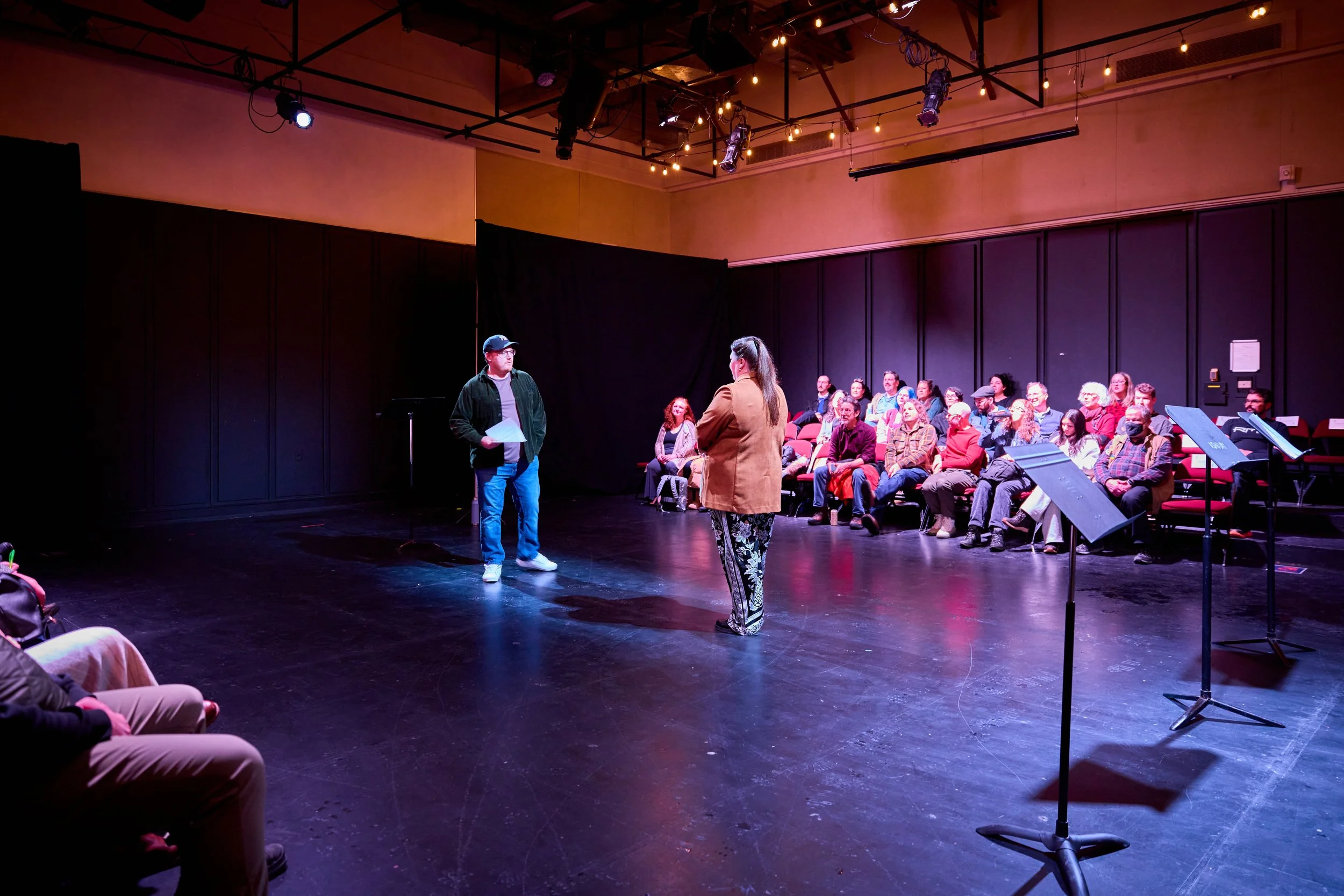 Une scène de théâtre ou d'audition avec deux personnes en avant, l'une tenant des feuilles et l'autre écoutant, entourées d'un public assis en demi-cercle dans une salle sombre, éclairée par des lumières sur le plafond.