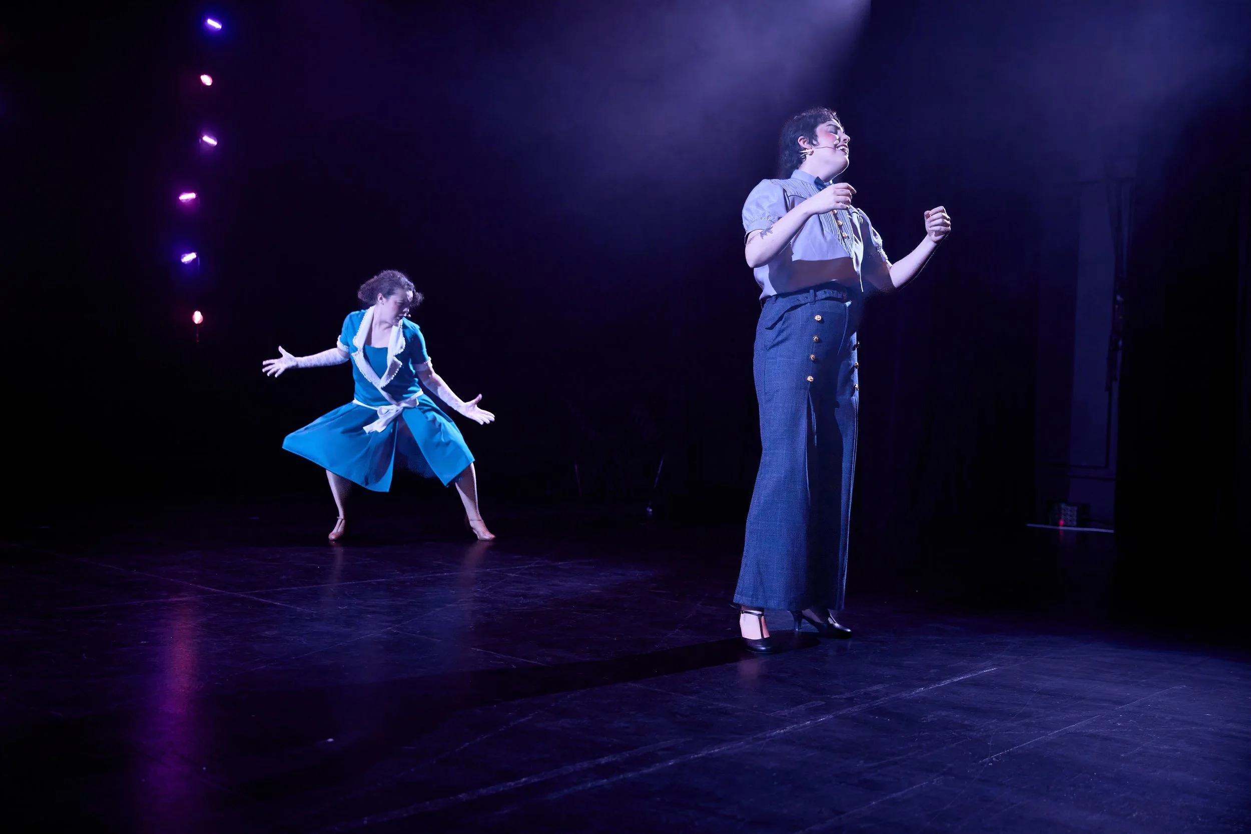 Deux personnes dans une scène de théâtre, une jeune fille en robe bleue et une femme en pantalon à motifs, sous un éclairage violet.