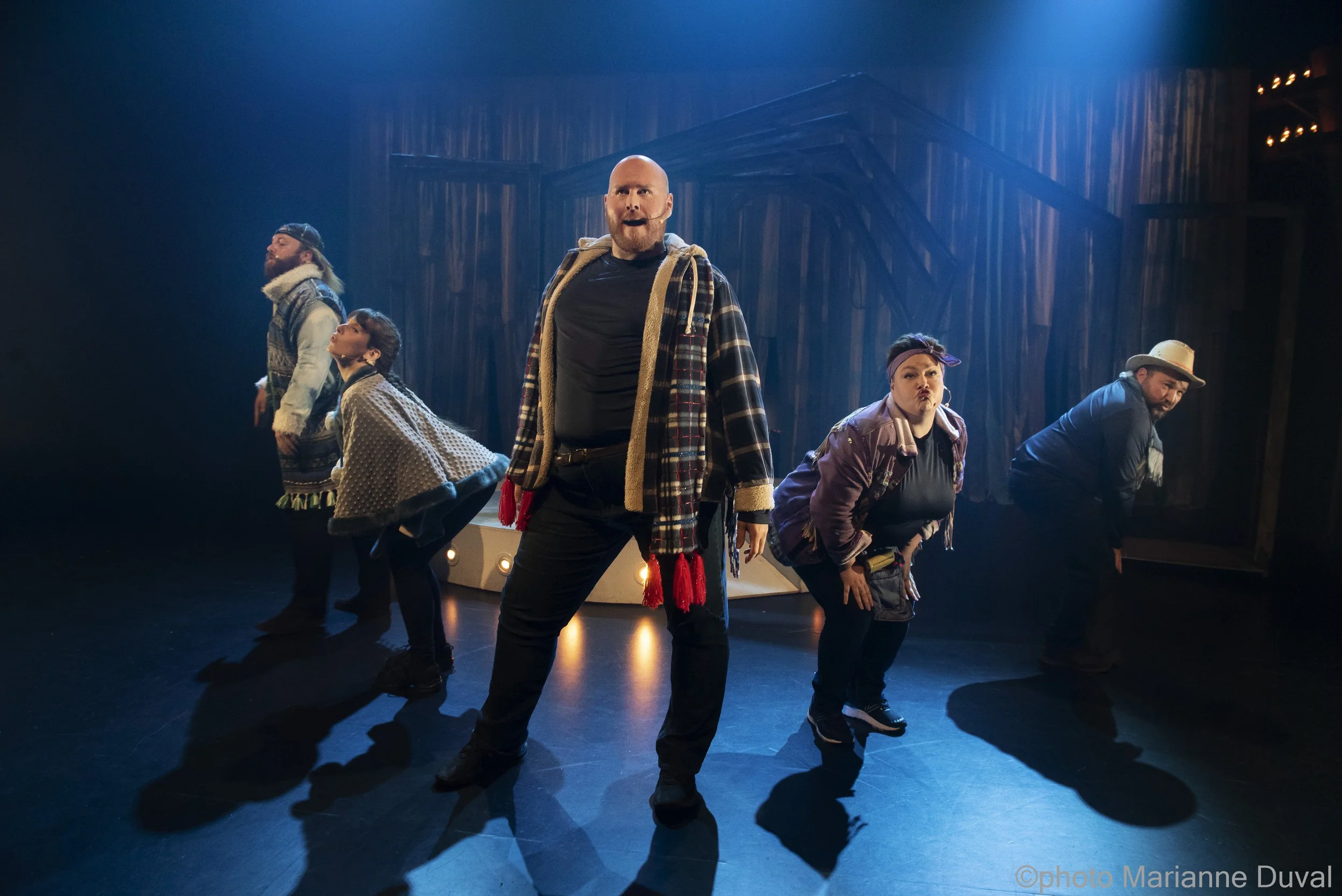 Un groupe de cinq personnes en costume de théâtre, posant sur scène avec un décor en bois en arrière-plan, sous un éclairage dramatique.