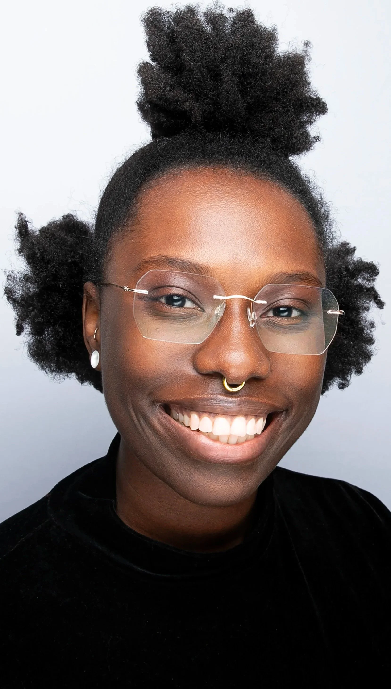 Canda-Leigh. Jeune femme africaine souriante avec des cheveux afro, portant des lunettes transparentes, une boucle d'oreille en perle, un anneau nasal en or, et un haut noir, devant un fond blanc.