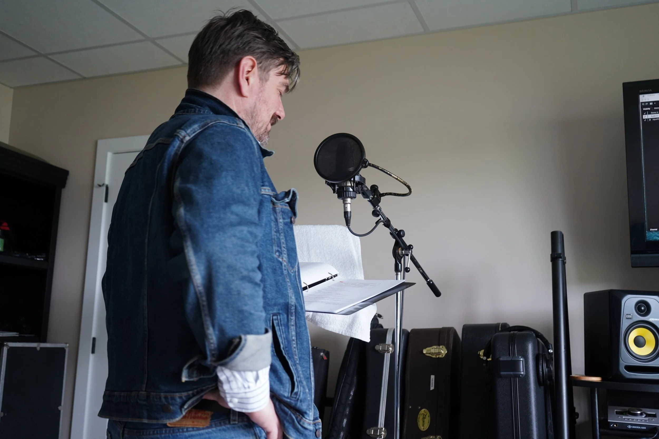 Un homme avec des cheveux bruns portant une veste en jean, tenant un casque audio ou un micro, se trouve dans une pièce avec des équipements audio et vidéo.