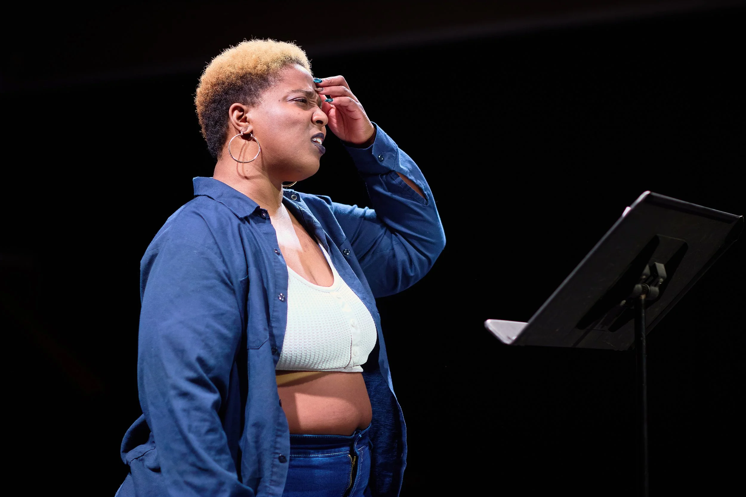 Une femme avec des cheveux courts blond foncé, portant une veste bleue, un t-shirt blanc, et de grands anneaux, semble stressée ou confuse devant une feuille de musique ou script sur un pupitre.