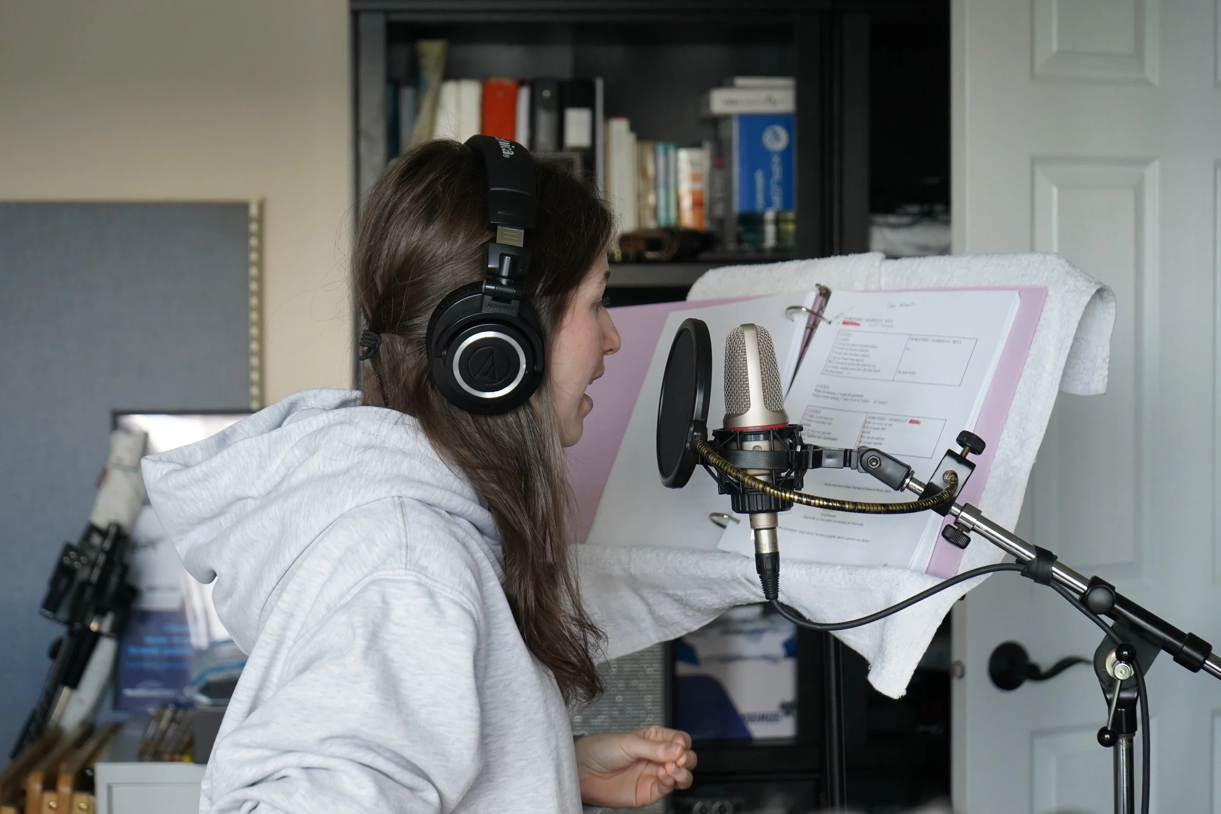 Une femme en train d'enregistrer une voix dans un studio à domicile, portant des écouteurs et parlant dans un microphone, avec une feuille de papier contenant des notes ou un script.
