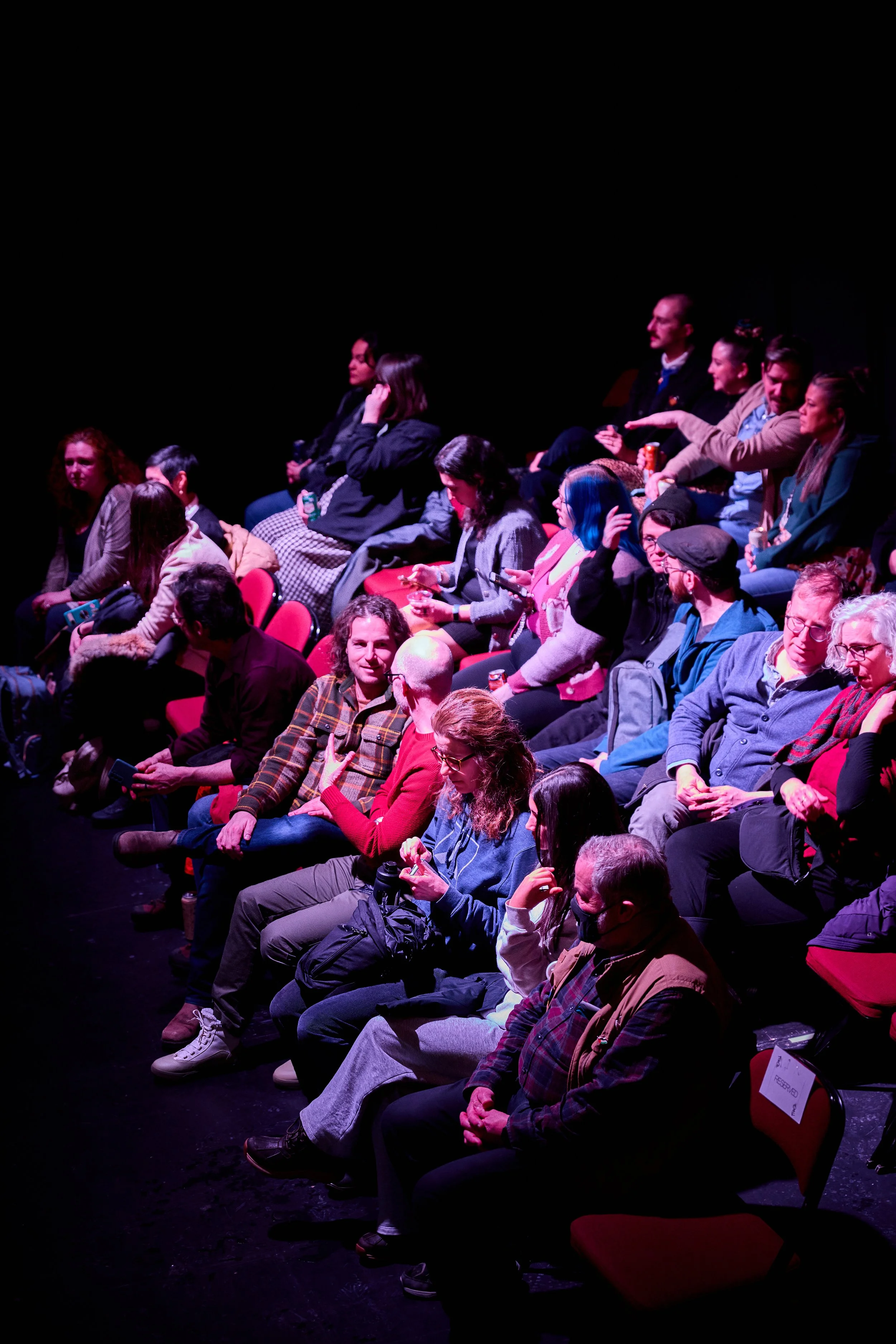 Public assis en auditorium sombre, certains utilisant leur téléphone, d'autres discutant ou attendant le début d'une présentation.