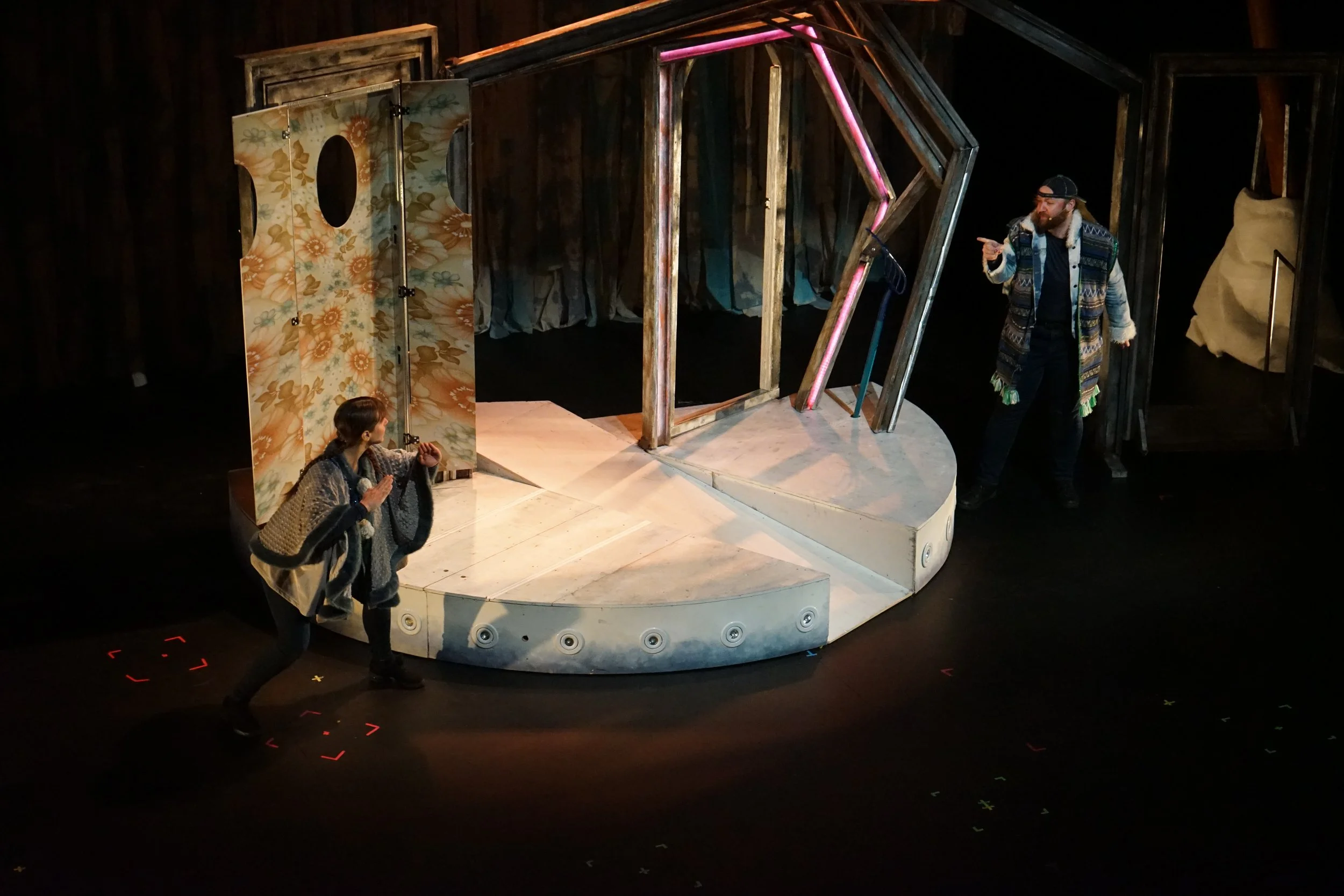 Deux acteurs en costume sur une scène de théâtre, un garçon à gauche et un homme à droite, avec un décor de maison en arrière-plan.