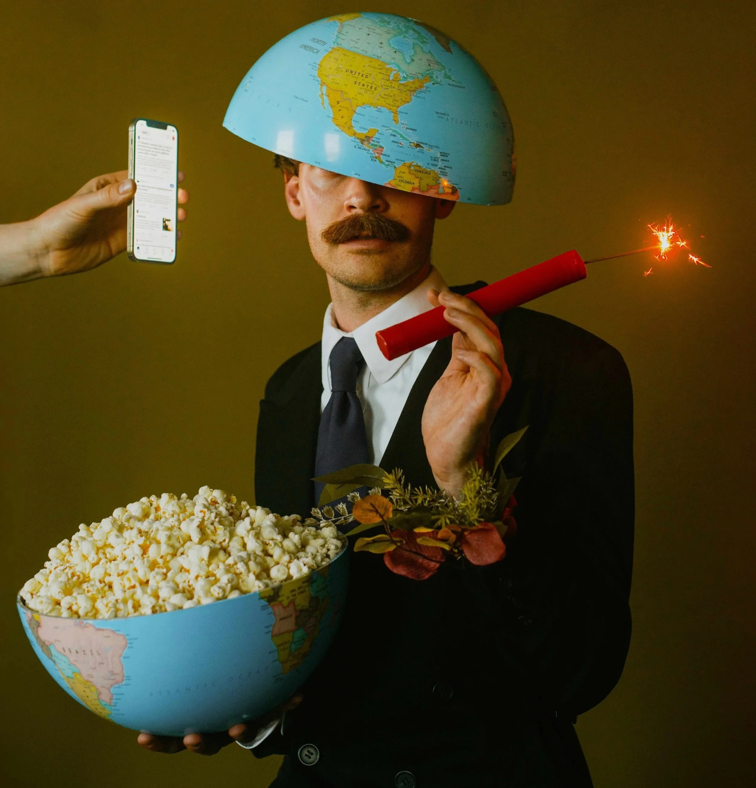 Un homme porte un globe terrestre sur la tête, tenant une pièce de monnaie et une étincelle de fusée, avec une boîte de popcorn dans l'autre main.