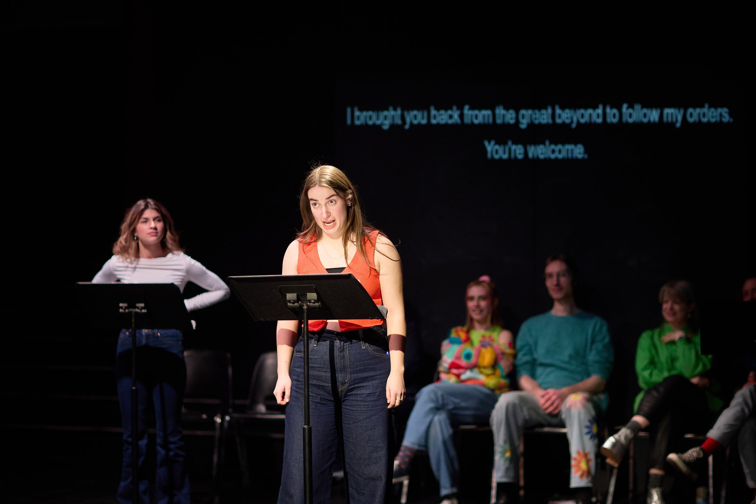 Une jeune femme parle devant un micro sur scène avec d'autres personnes assises en arrière-plan et un texte projeté à l'écran derrière elle.