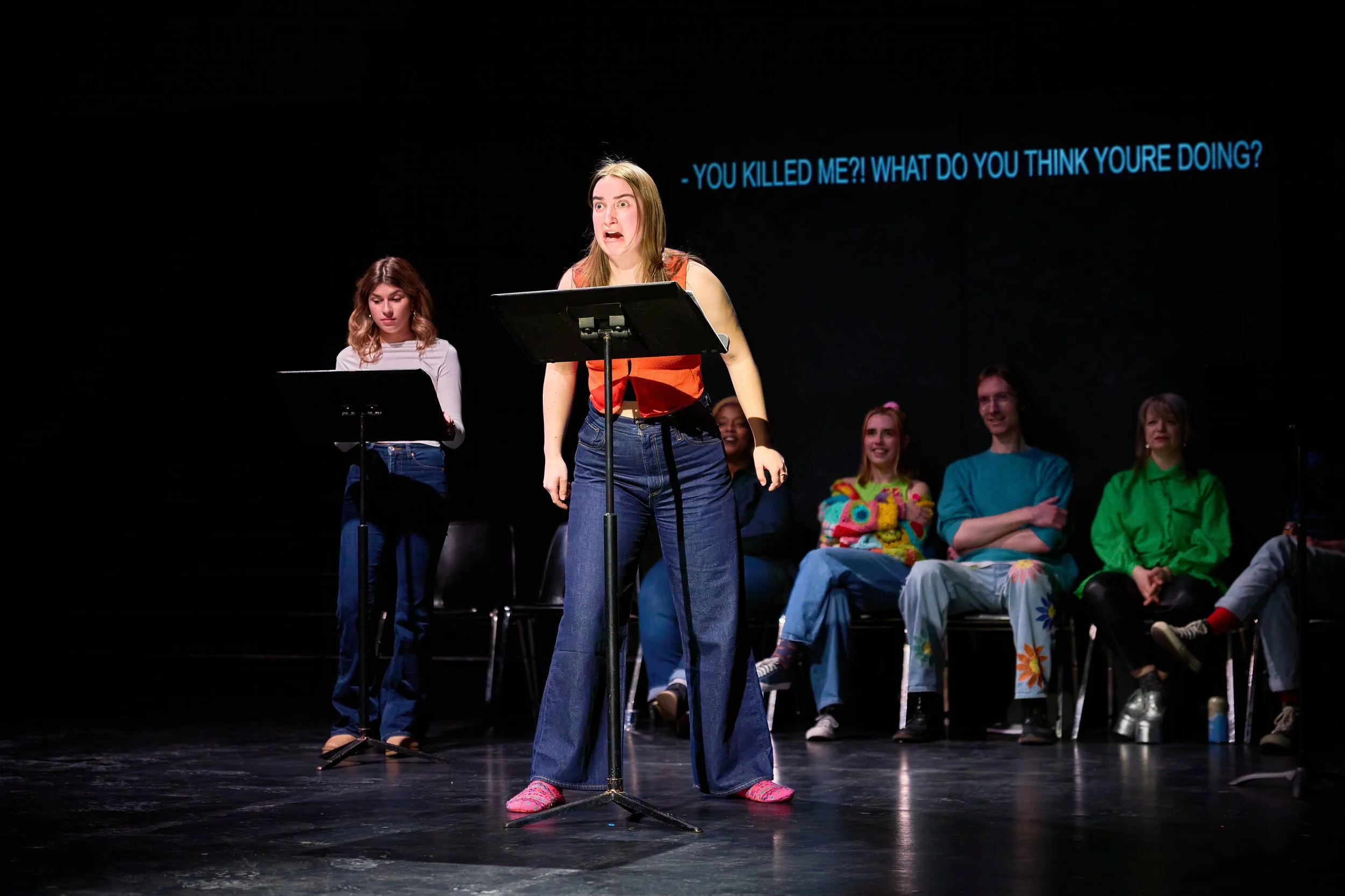 Une scène de théâtre avec des jeunes actrices, dont une en première plan qui exprime une grande émotion. En arrière-plan, d'autres jeunes assises et regardant l'actrice principale, avec des sous-titres en anglais disant: 'You killed me?! What do you 