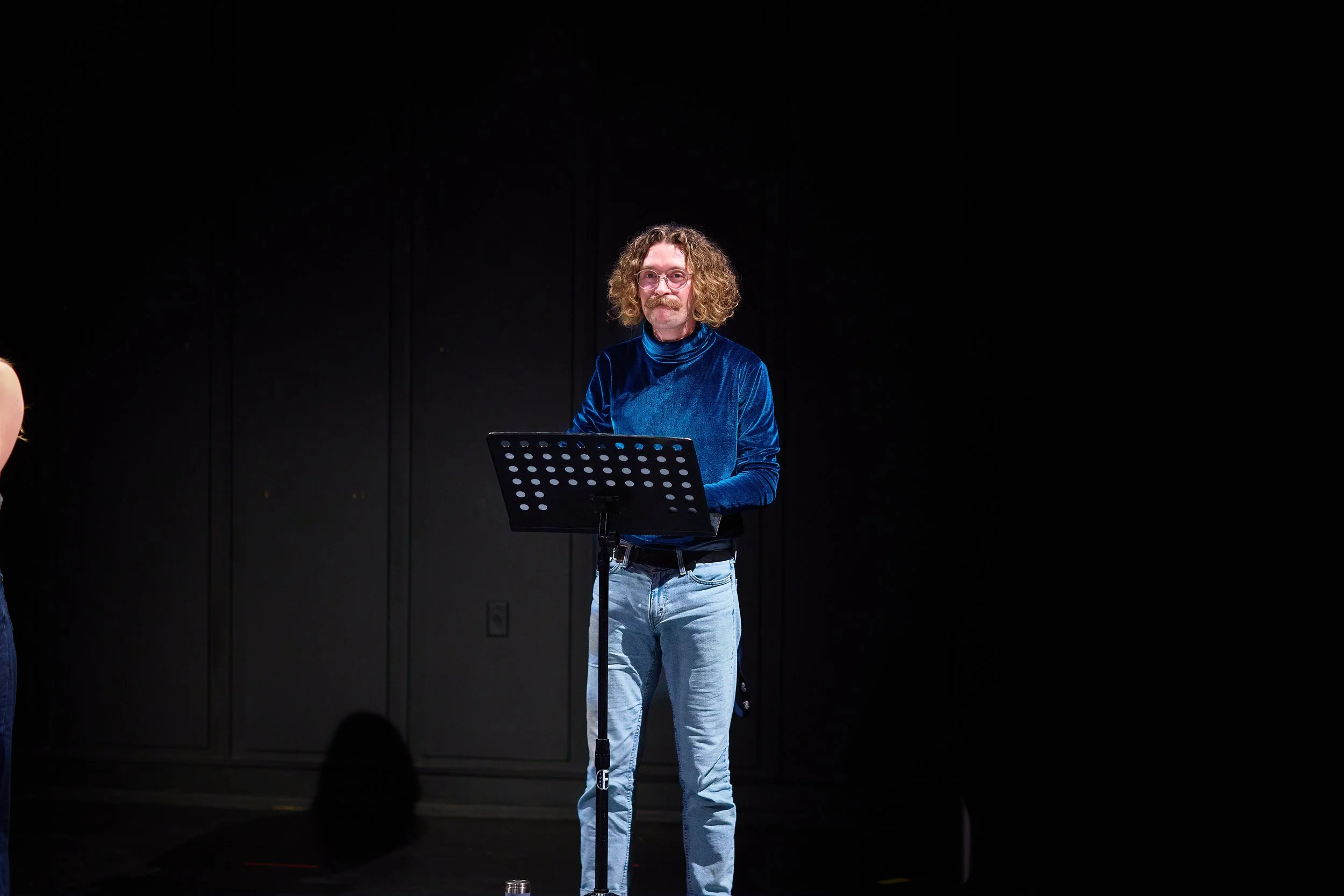 Homme avec cheveux bouclés et moustache, portant un haut en velours bleu et des jeans, se tenant devant un pupitre noir dans une scène sombre.