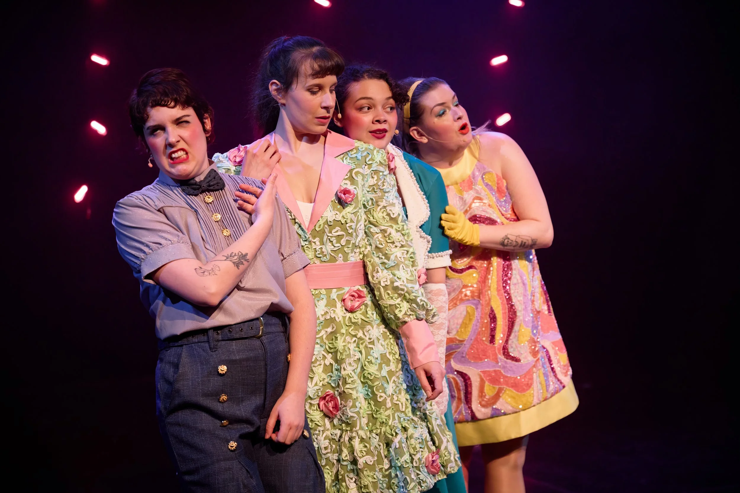 Cinq femmes en costume théâtral sur scène, faisant une expression dramatique, avec un fond noir et des lumières roses.
