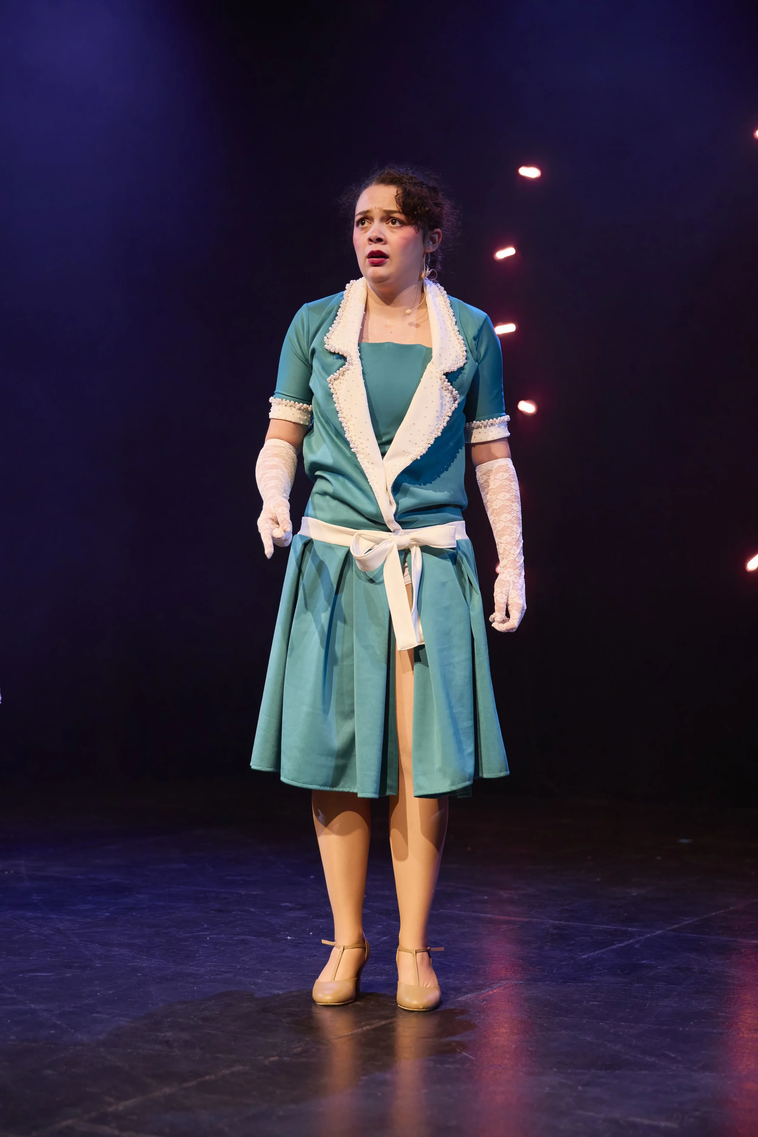 Une femme dans un costume de scène, exprimant une émotion de surprise ou d'inquiétude, sur scène avec un fond sombre et des lumières.