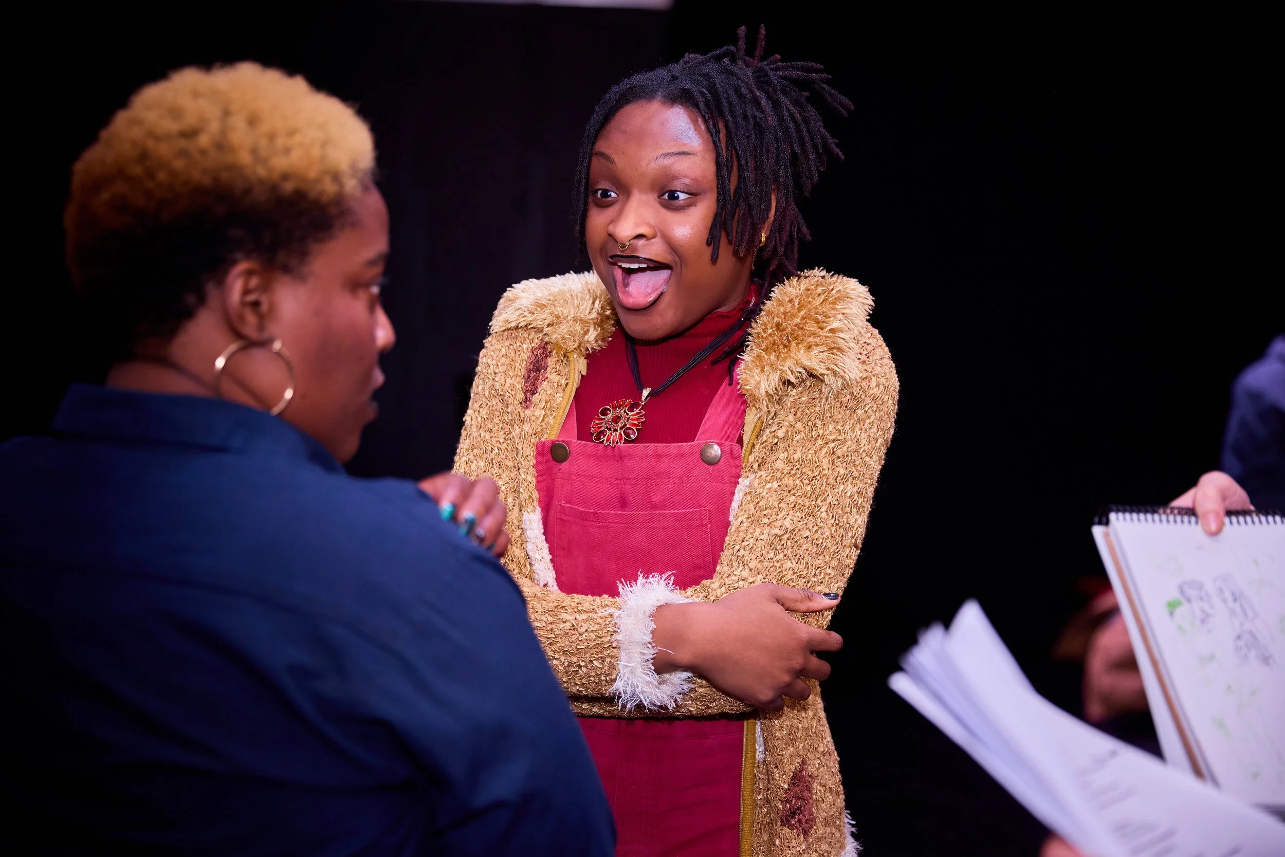 Deux femmes discutent, une d'elles a un sourire enthousiaste et un tatouage au visage, l'autre est vu de dos, tenant un carnet avec des dessins.