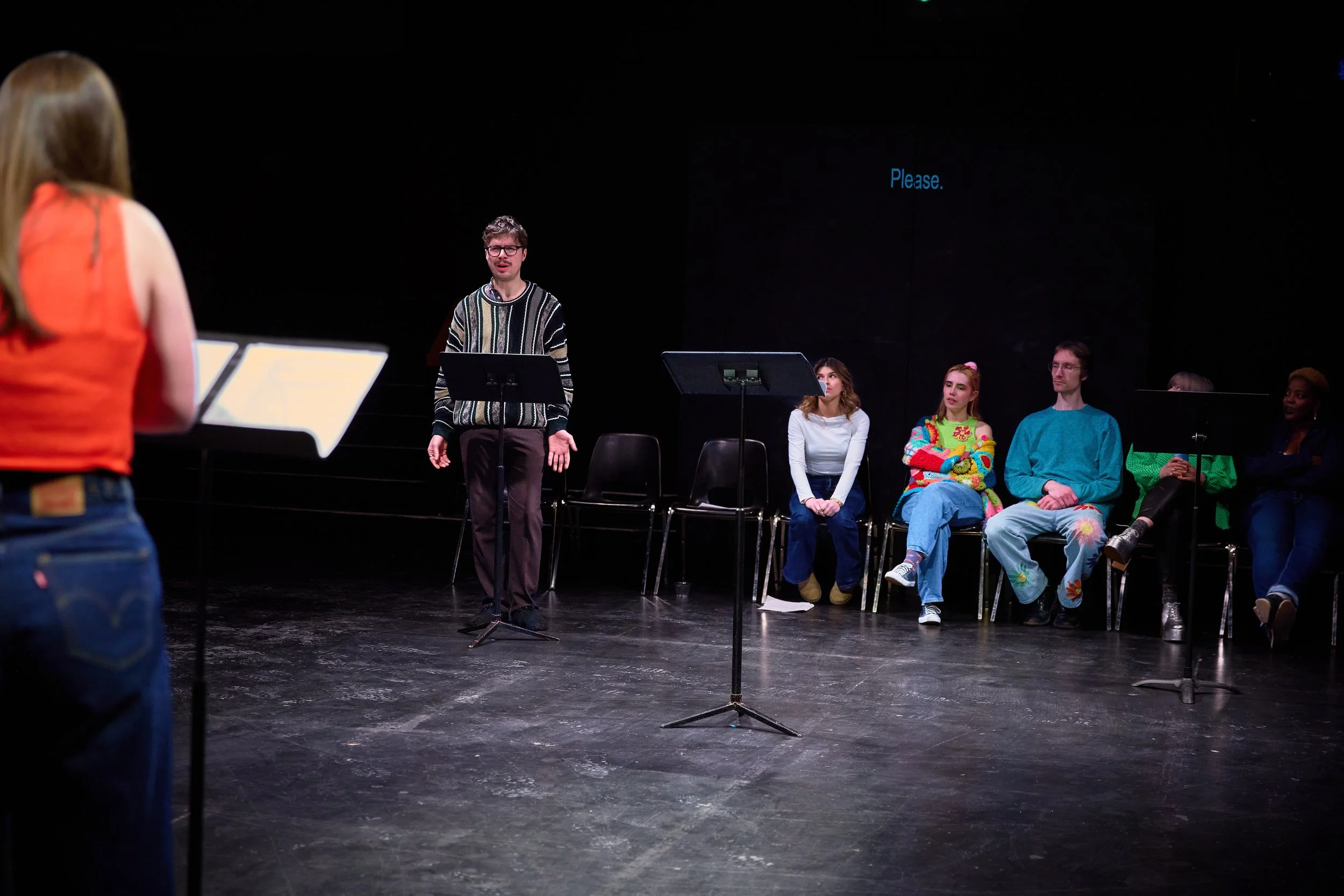 Jeune homme en train de parler devant un groupe de personnes assises sur scène, avec un fond noir et un écran affichant le mot 'Please'.