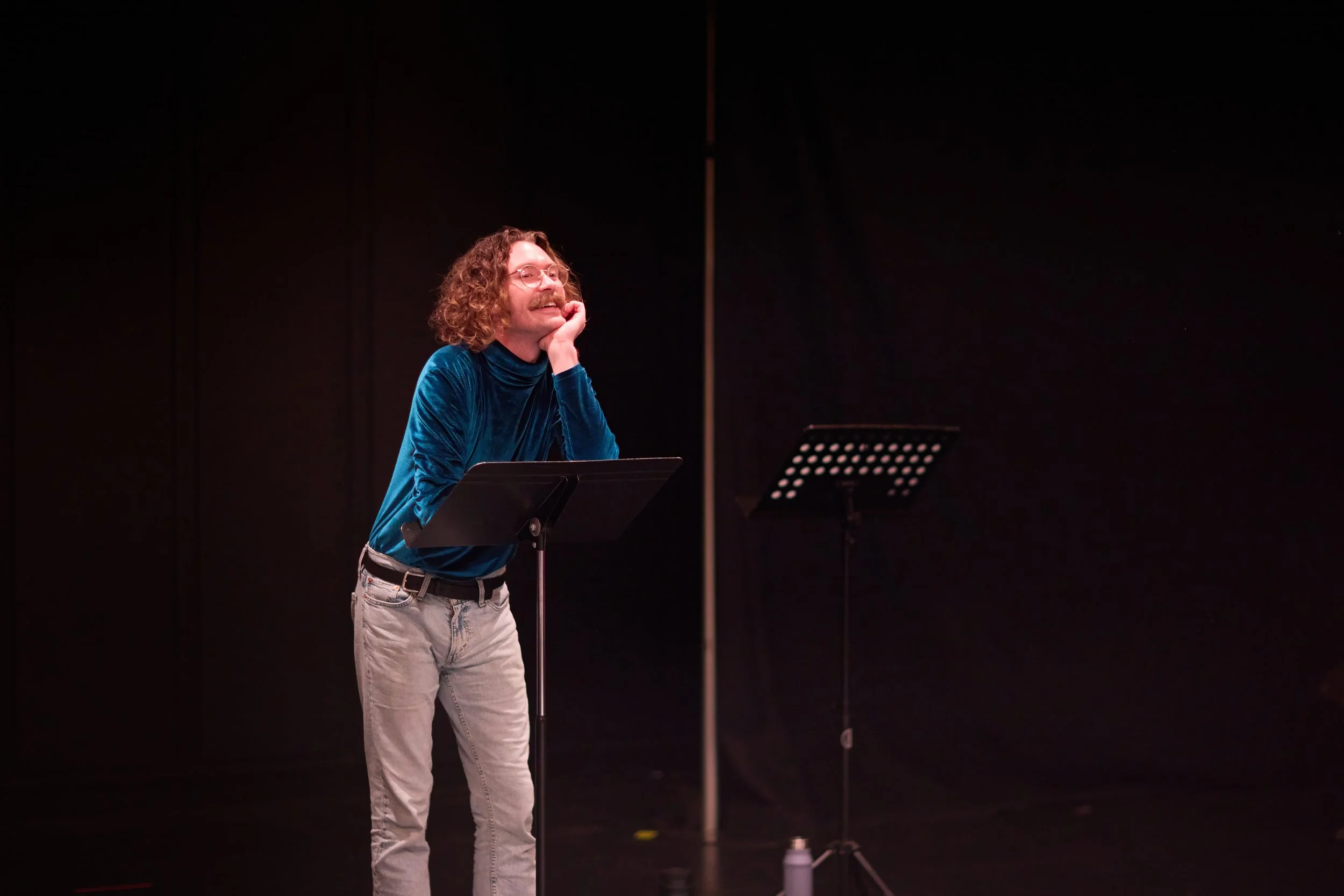 Un homme aux cheveux longs et bouclés, portant des lunettes et une tenue bleue, sourit et regarde vers le haut dans un studio avec un pupitre et un microphone.