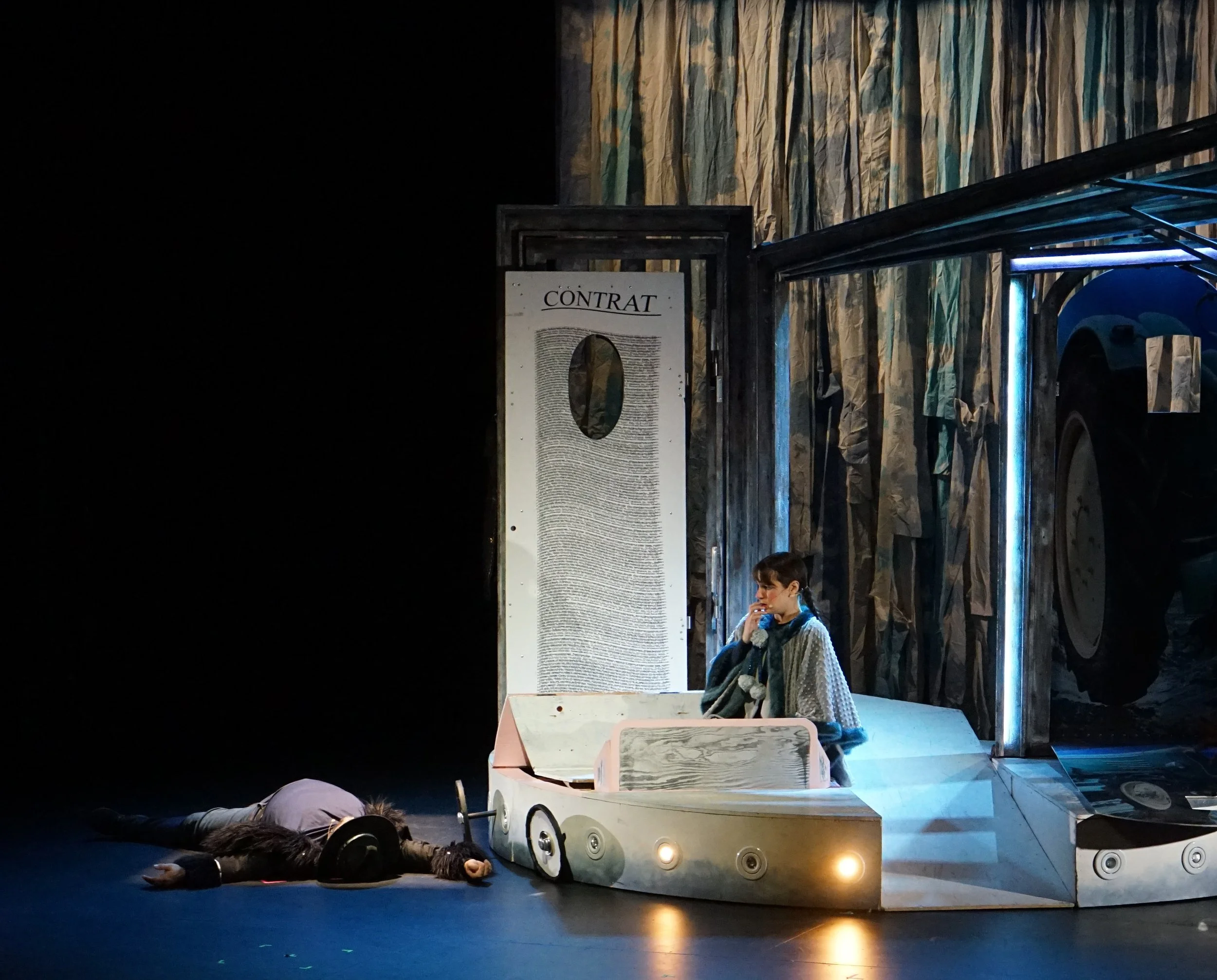 Scène de théâtre avec deux acteurs, un allongé sur le sol et l'autre assis à une table, dans un décor de scène avec un panneau marqué 'CONTRAT' et une grande roue ou machine.