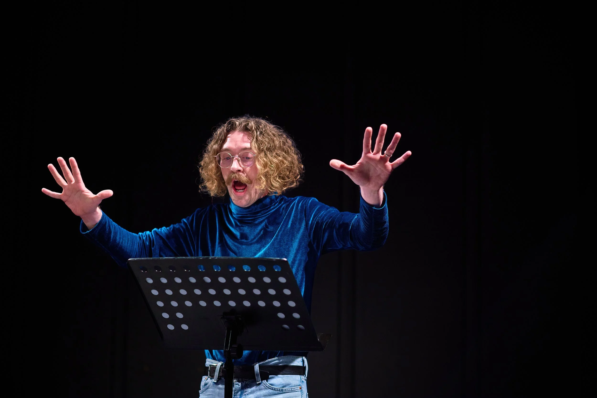 Un homme aux cheveux frisés longs, portant des lunettes rondes, une veste en velours bleu et un jean, semble chanter ou parler avec expressivité, avec un pupitre en métal noir avec des trous devant lui, sur fond noir.