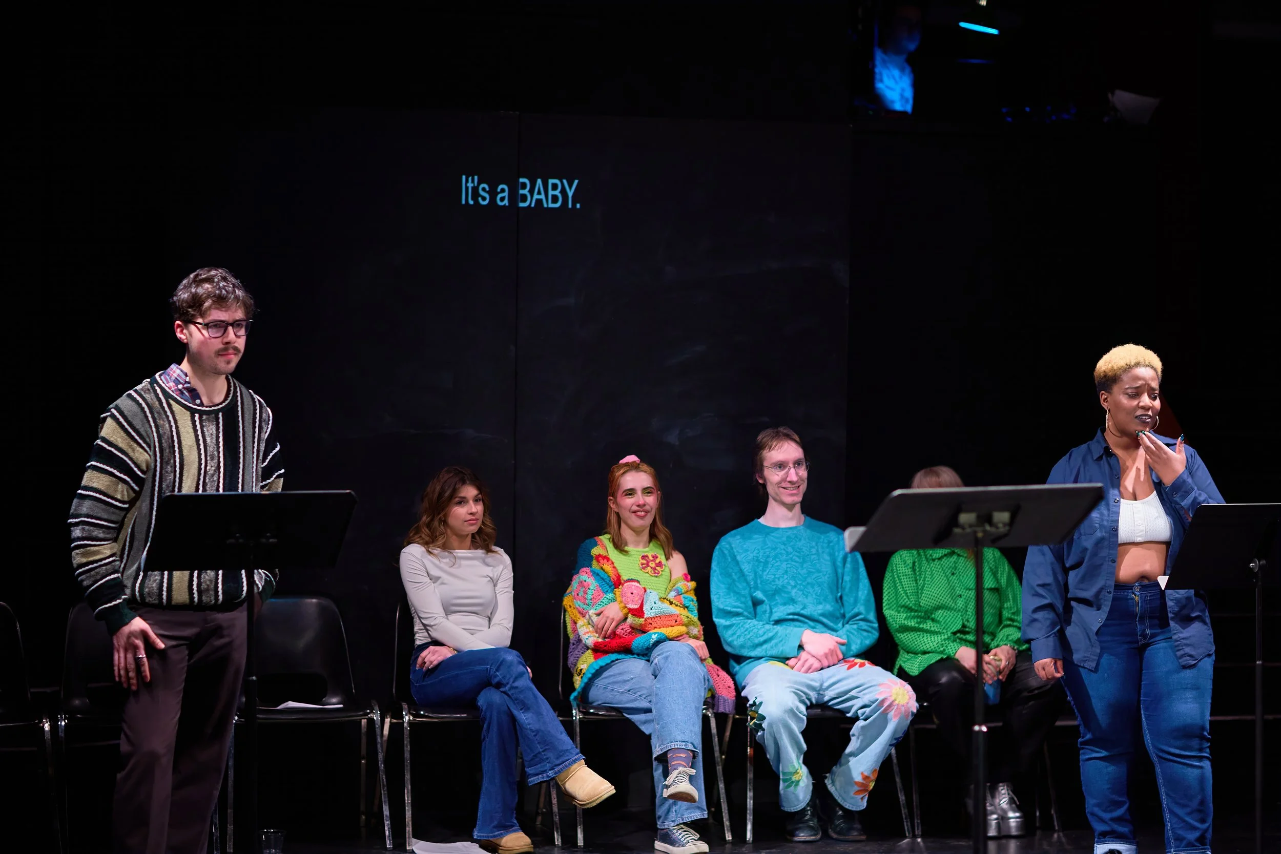 Un groupe de personnes assises sur des chaises sur scène lors d'une représentation théâtrale, avec une femme debout parlant, et un fond noir avec le texte "It's a BABY".
