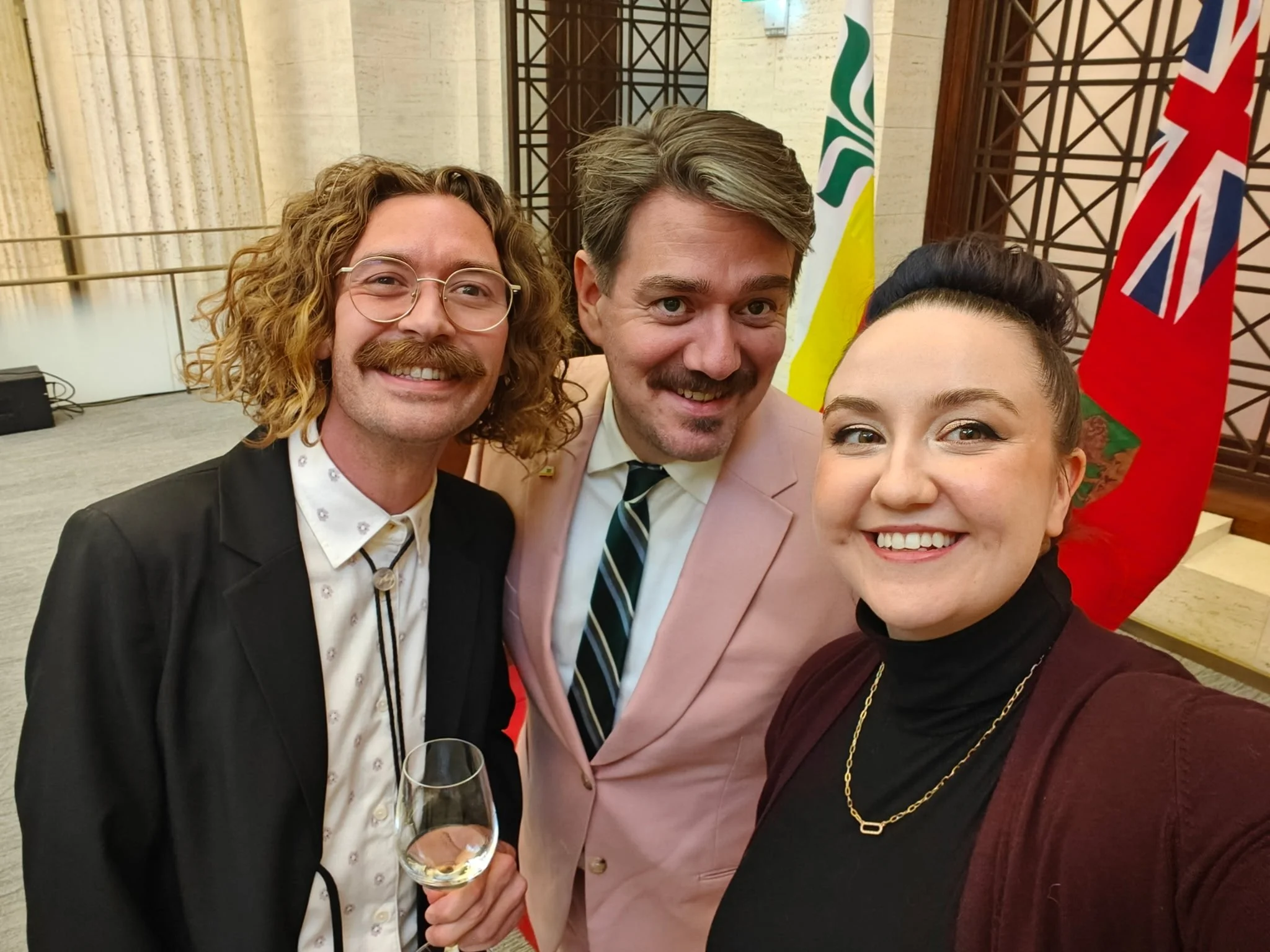Trois personnes souriantes, un homme avec des lunettes et une moustache, une femme avec un chignon, et un homme avec une moustache, posant pour une selfie, avec des drapeaux au fond.