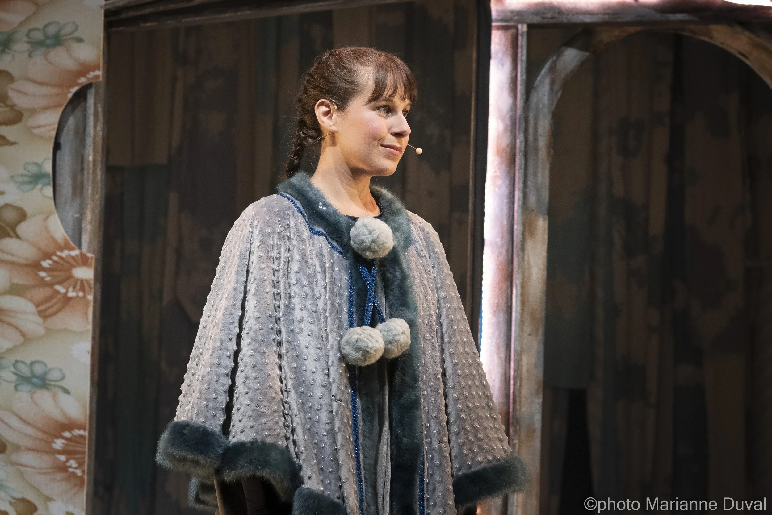 Une femme dans un costume théâtral, portant un manteau gris avec des détails en fourrure et des pompons, se tient sur scène avec un fond en bois.