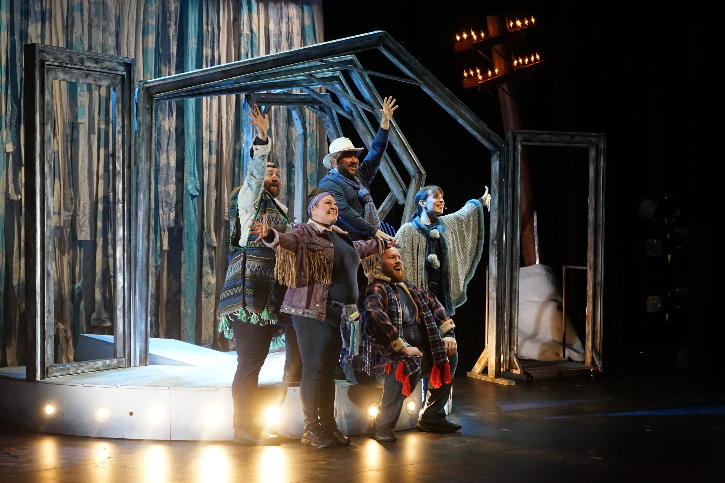 Groupe de six acteurs en costume sur scène de théâtre, posant joyeusement, avec décor en bois et éclairages chaleureux.