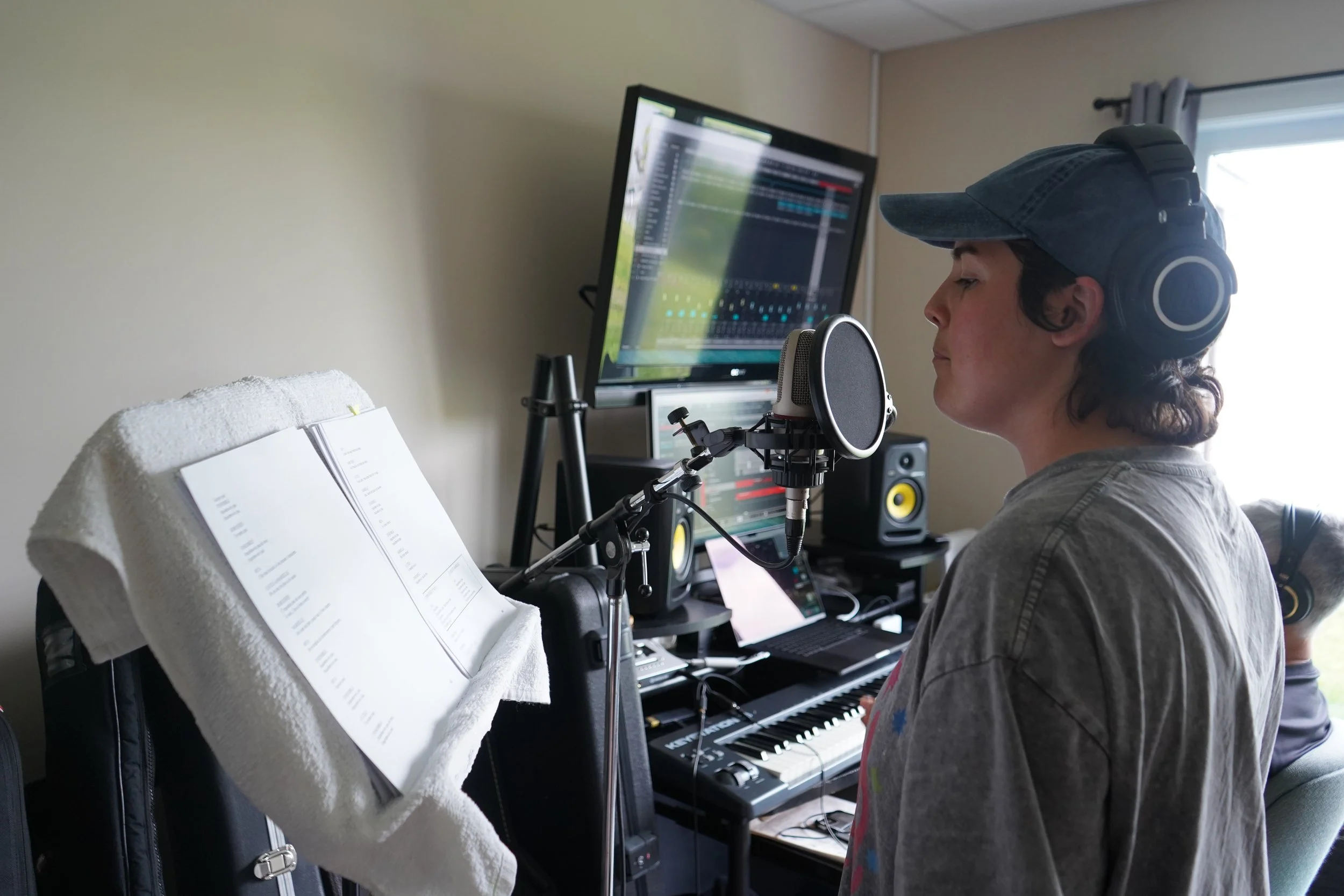 Une personne avec un casque et des écouteurs en studio d'enregistrement, devant un microphone, avec un écran d'ordinateur et un étui de guitare ou de clavier en arrière-plan.