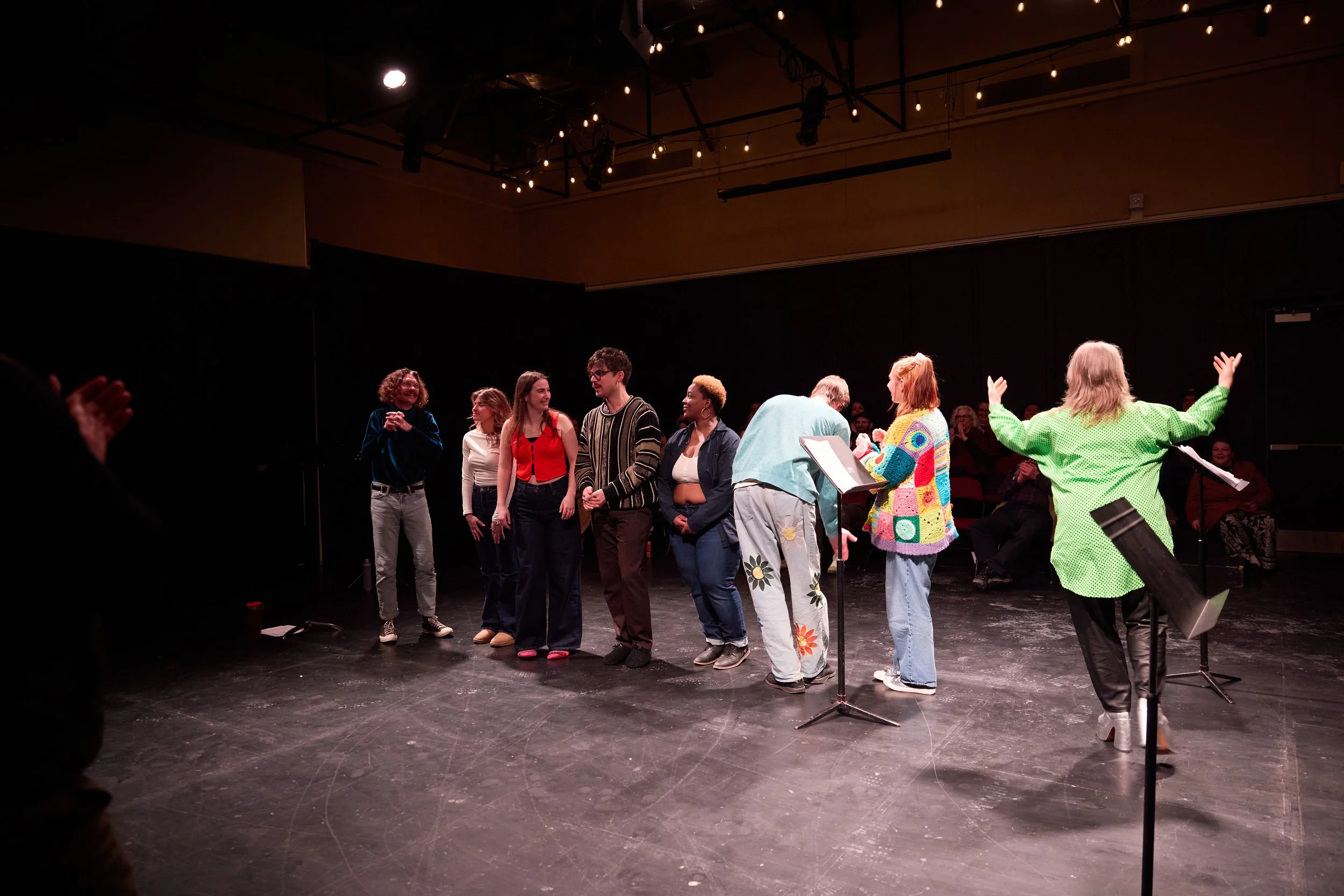 Groupe de personnes debout sur scène lors d'une performance, applaudissant et interagissant, avec un public en arrière-plan, dans une salle de théâtre.