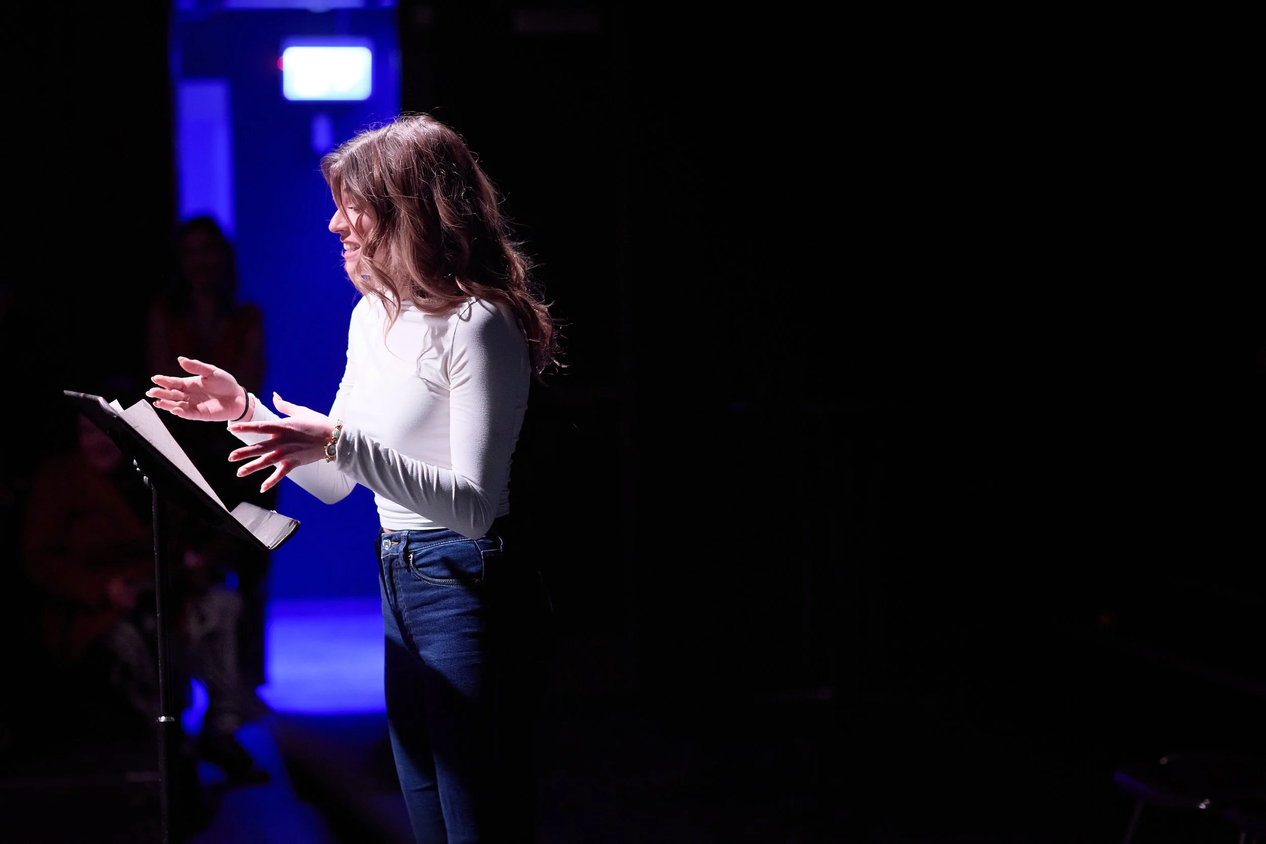 Une femme avec des cheveux bruns bouclés, portant un haut blanc et un jean, parle ou chante avec un micro en main, sur scène.