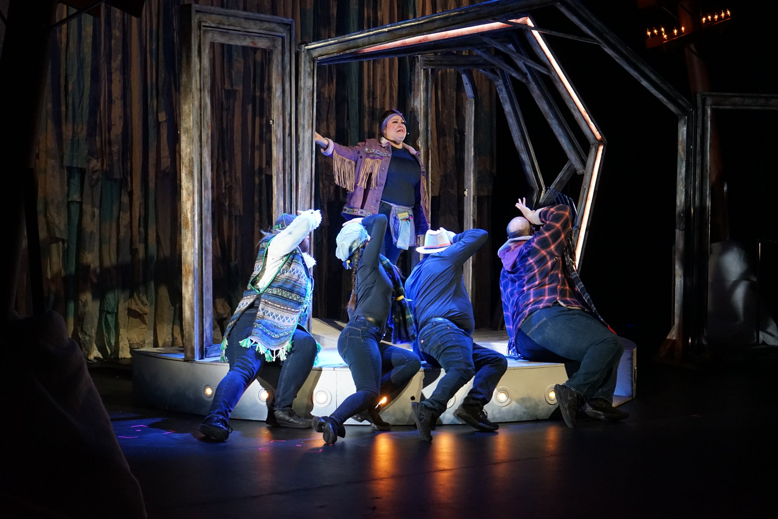 Une scène de théâtre avec une actrice sur scène, entourée de quatre personnes en costume, dans un décor en bois rustique, à l'intérieur d'une salle de spectacle.