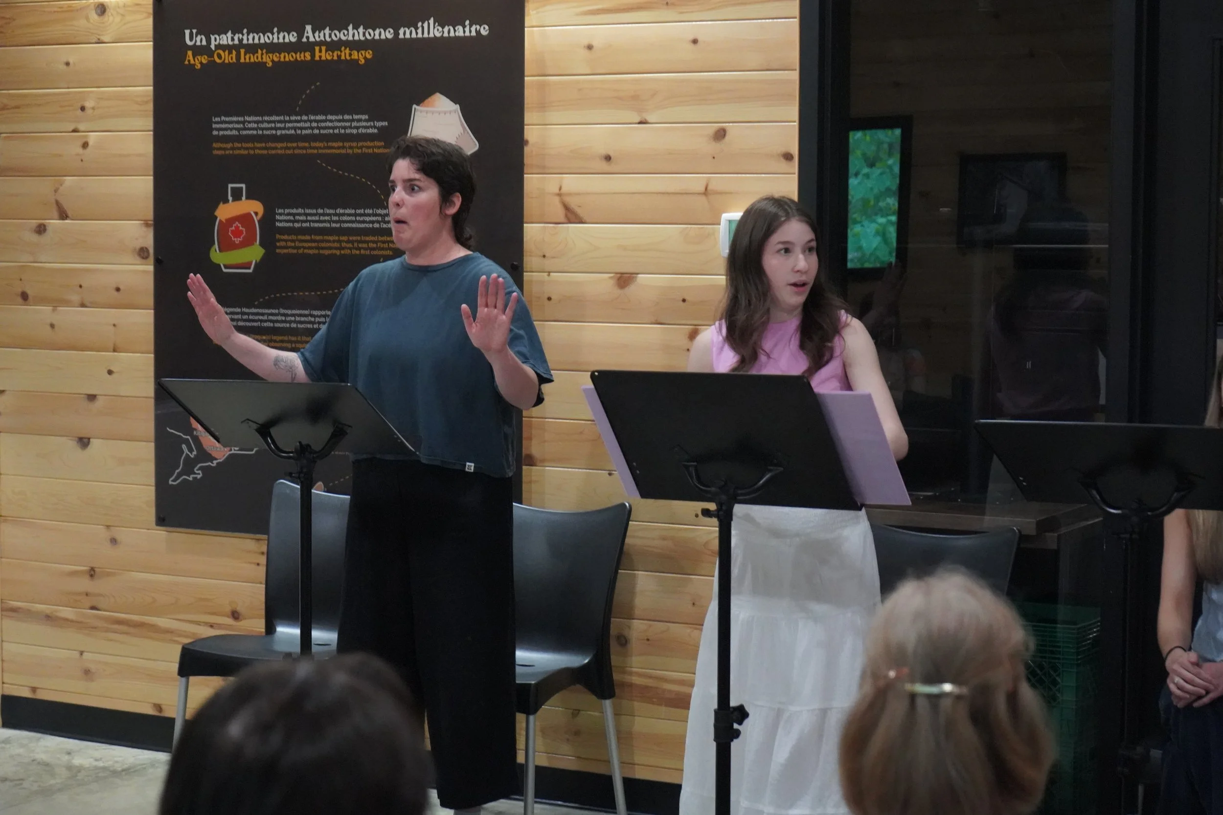 Deux jeunes femmes participent à une présentation ou un discours dans une pièce avec des murs en bois. La femme à gauche porte un t-shirt bleu et parle avec une gestuelle expressive, tandis que la femme à droite porte un haut rose clair et semble éco