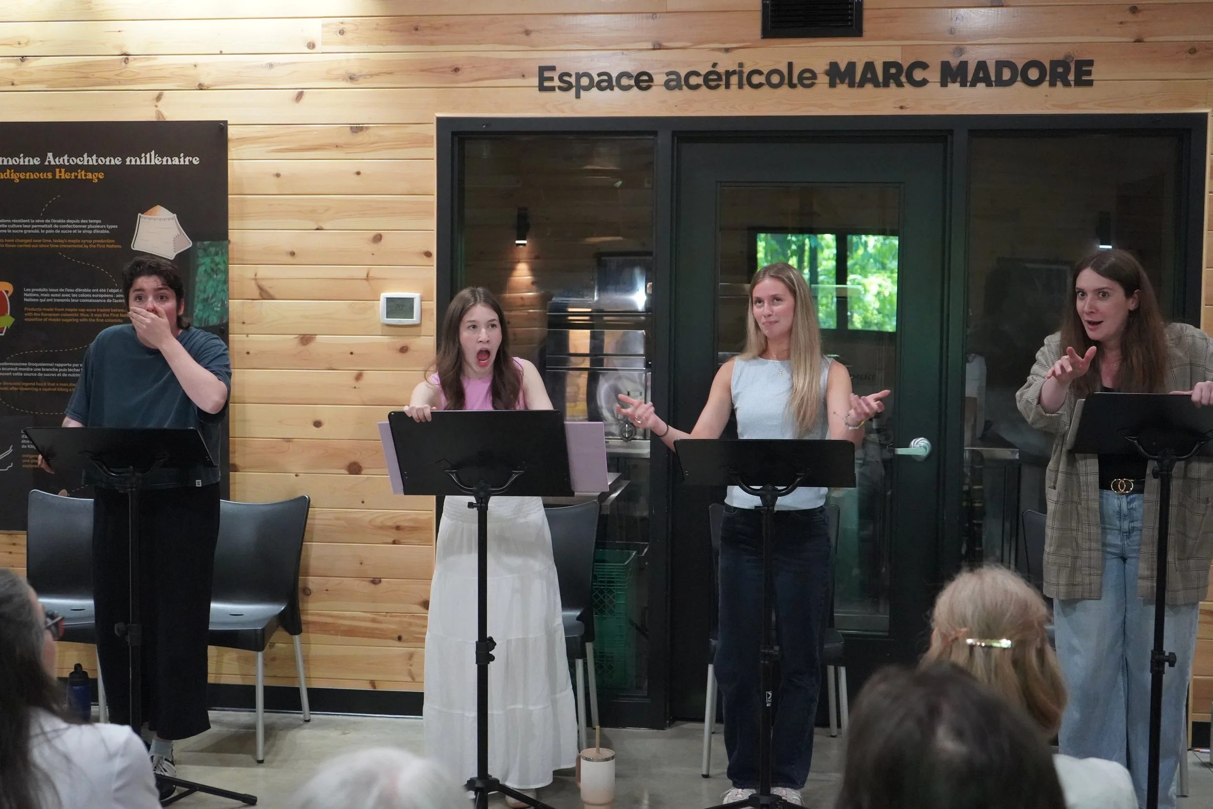 Quatre jeunes femmes se produisent comme choristes ou oratrices lors d'un spectacle, derrière des pupitres musicaux, dans une salle à bois avec fenêtres donnant sur une scène extérieure verdoyante.