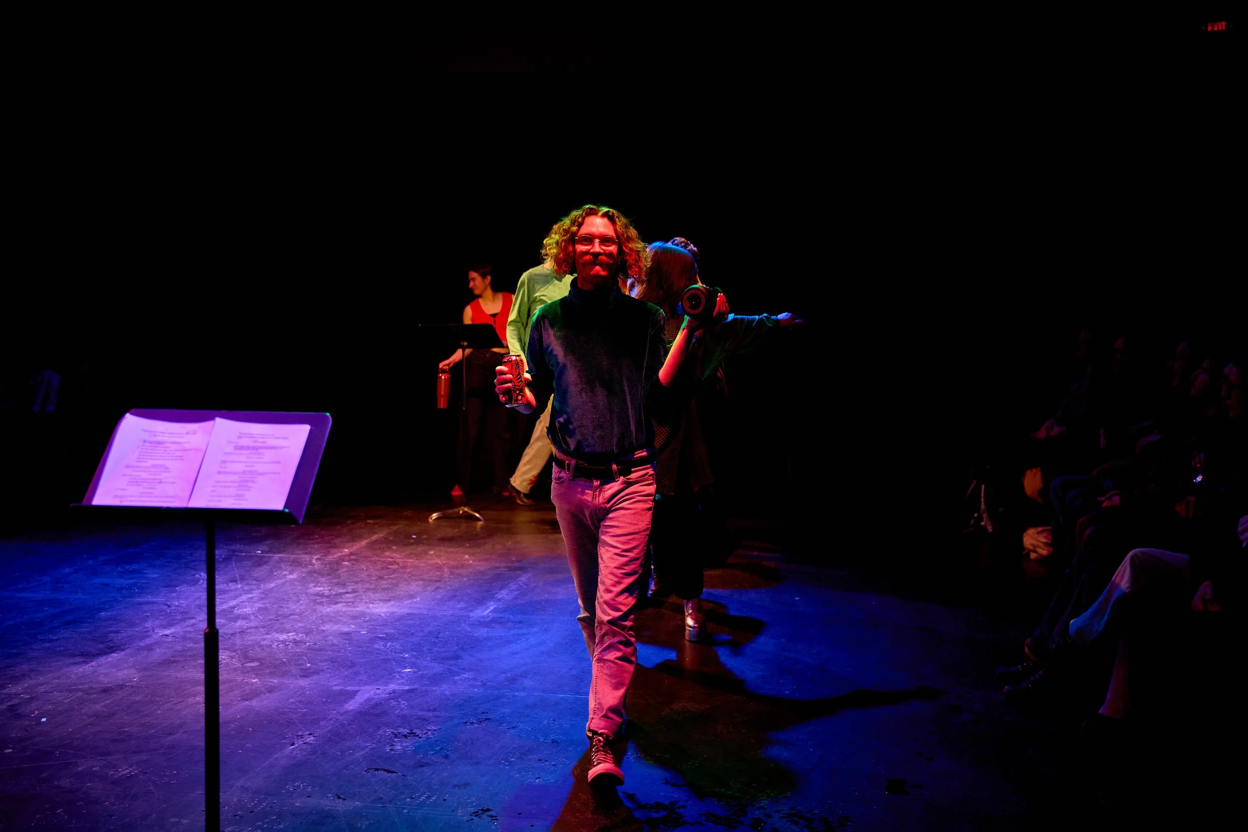 En scène, un homme avec des cheveux bouclés souriant, portant un t-shirt noir et un pantalon clair, marche vers l'avant avec d'autres personnes en arrière-plan. Il y a un pupitre avec une feuille de papier sur la gauche, dans une pièce sombre avec un
