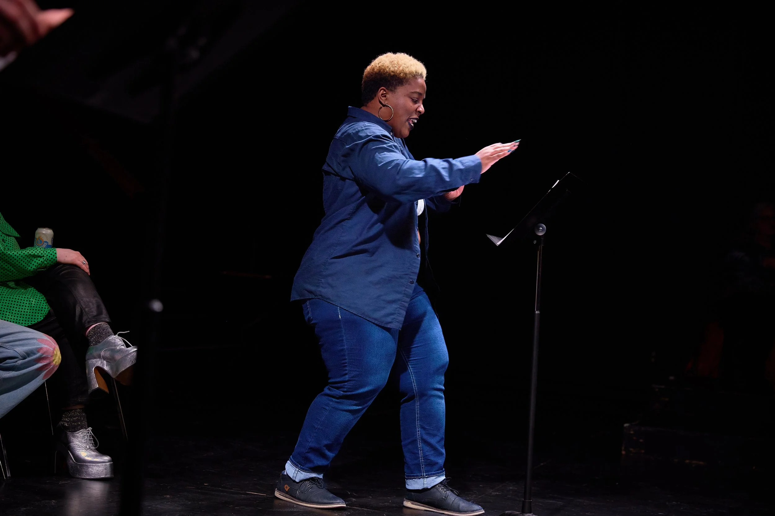 Une femme qui récite ou performe sur scène, debout devant un pupitre avec une musique ou un script, mais aucune audience visible.