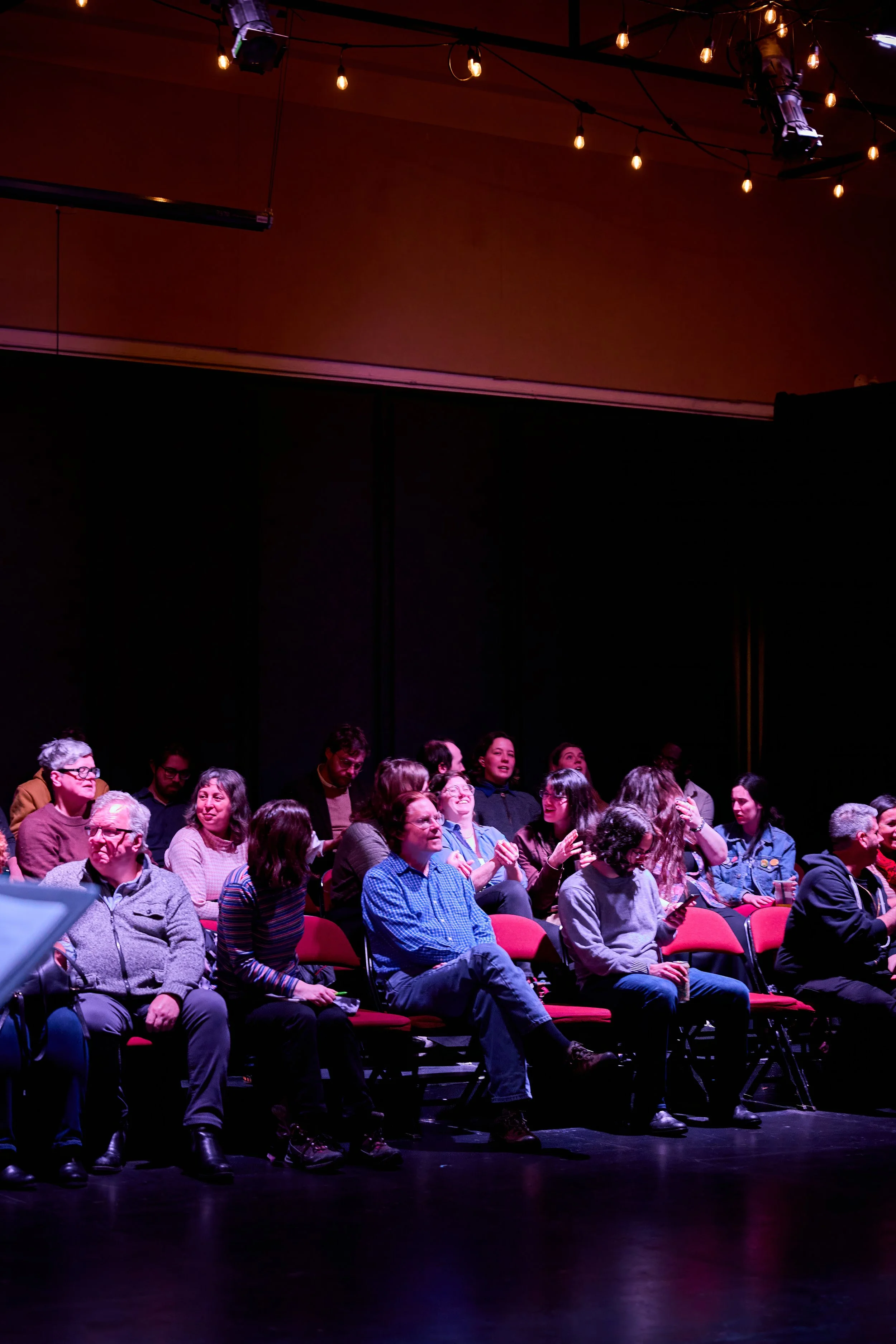 Groupe de personnes assises dans une salle sombre, en train de rire et de parler, avec un éclairage tamisé et des ampoules suspendues au plafond.