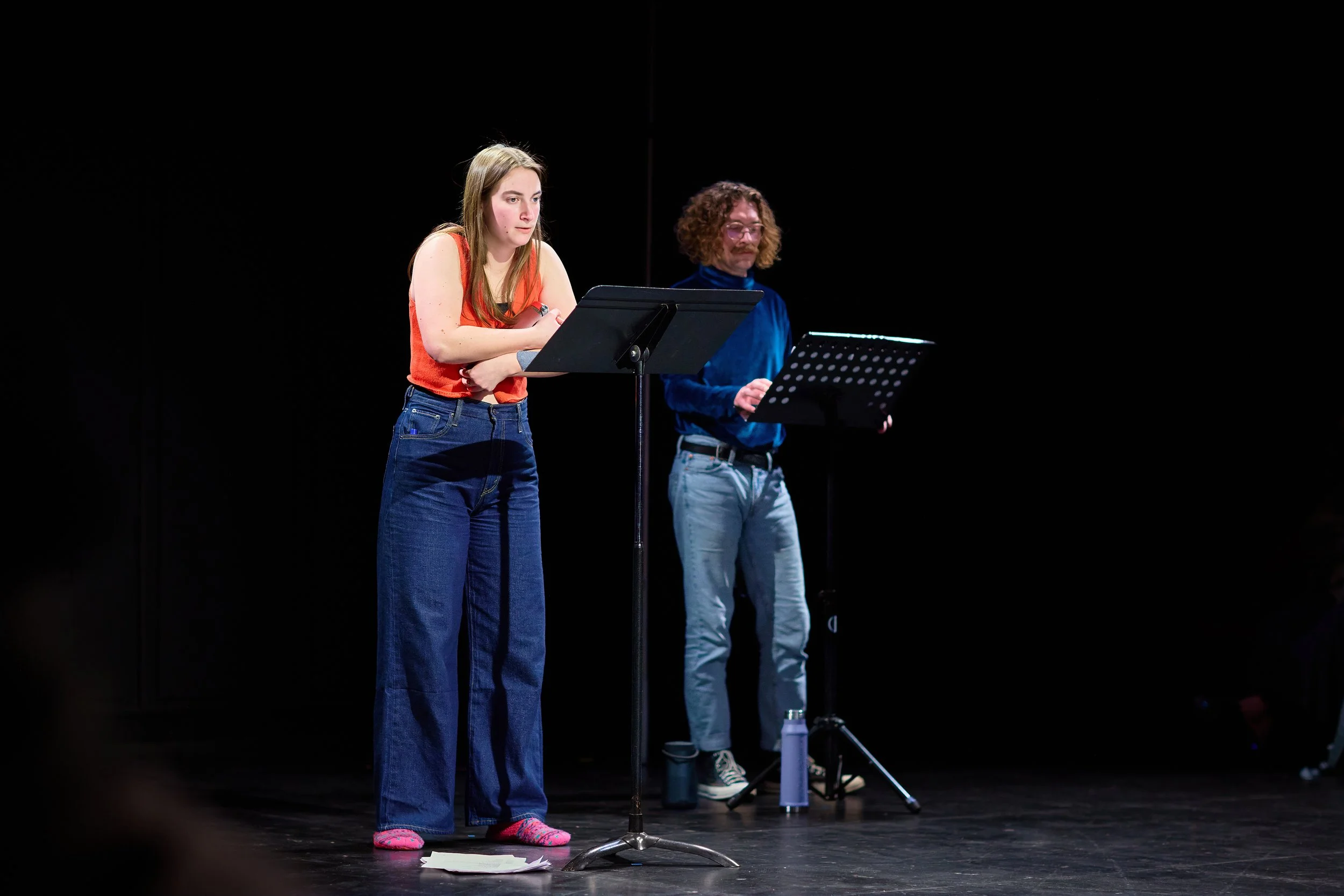 Deux personnes sur scène, une femme à gauche avec une expression concentrée, et un homme à droite avec des notes, en pleine lecture ou répétition, dans un espace sombre.