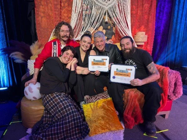 Créations In Vivo. Cinq personnes souriantes posent ensemble sur un canapé décoré de coussins colorés, tenant des certificats ou diplômes, dans un décor coloré avec des rideaux en arrière-plan.