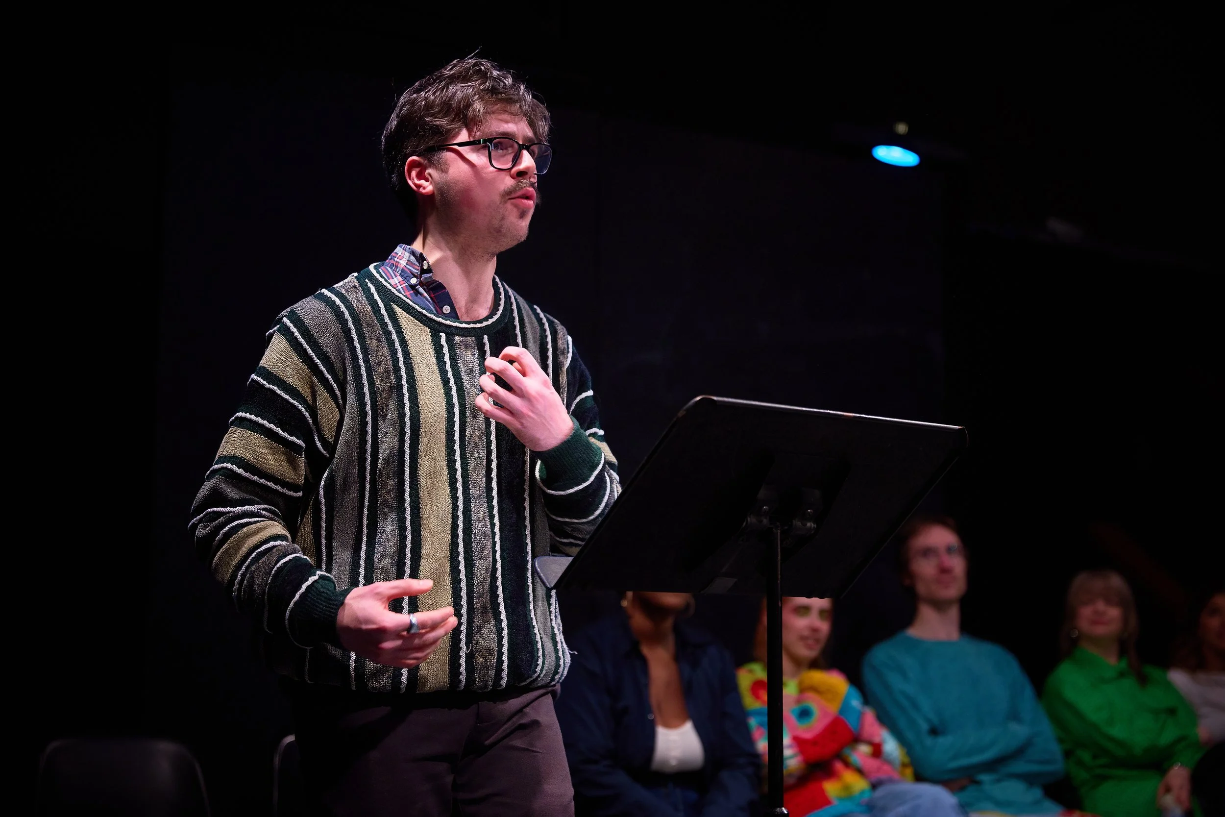 Un jeune homme avec des lunettes et un pull rayé parle devant un micro lors d'une présentation ou d'une pièce de théâtre, avec un groupe de personnes assises derrière lui dans un décor sombre.