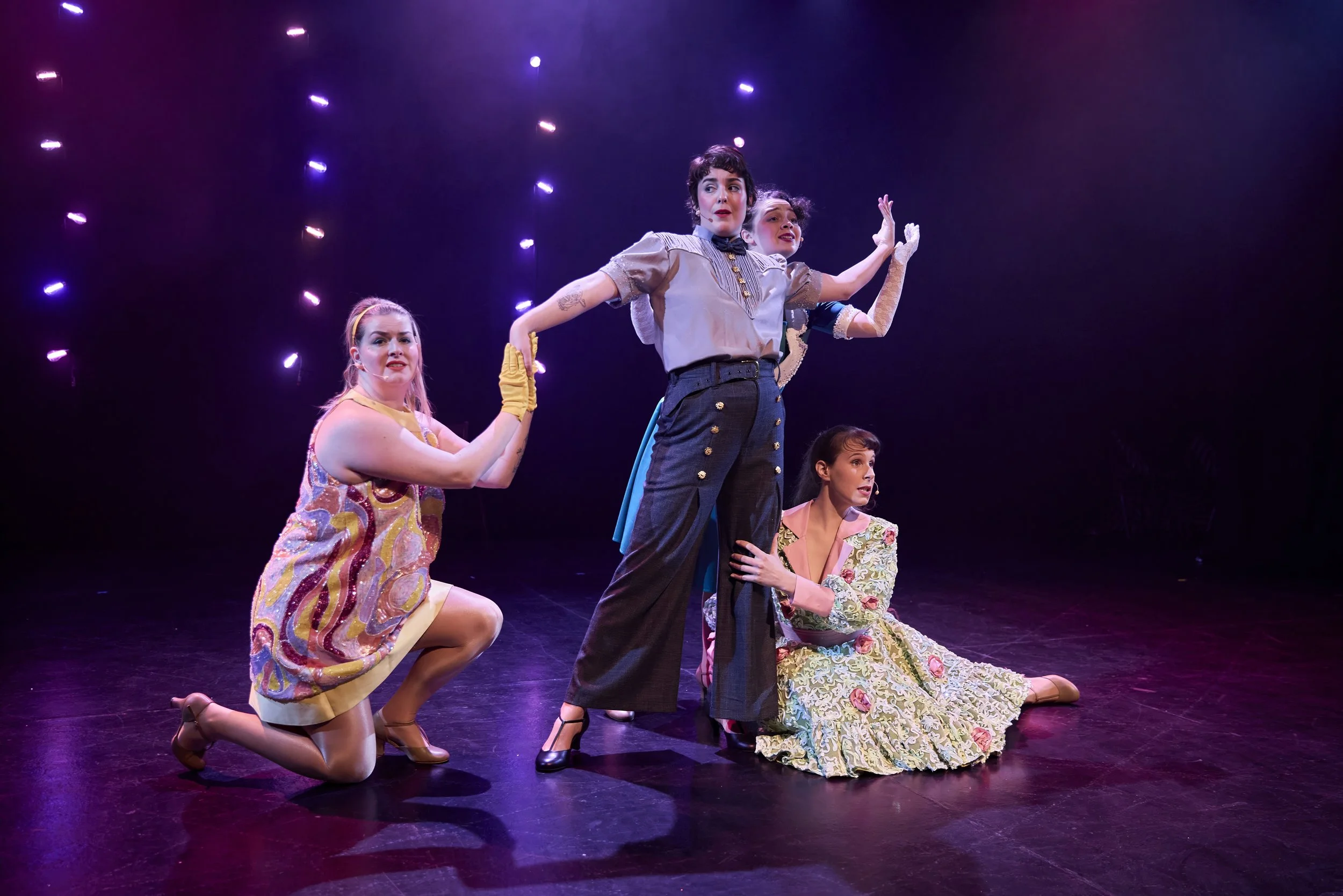 Créations In Vivo. Quatre femmes en costumes de théâtre sur scène avec lumières colorées derrière elles, posant pour une scène dramatique.