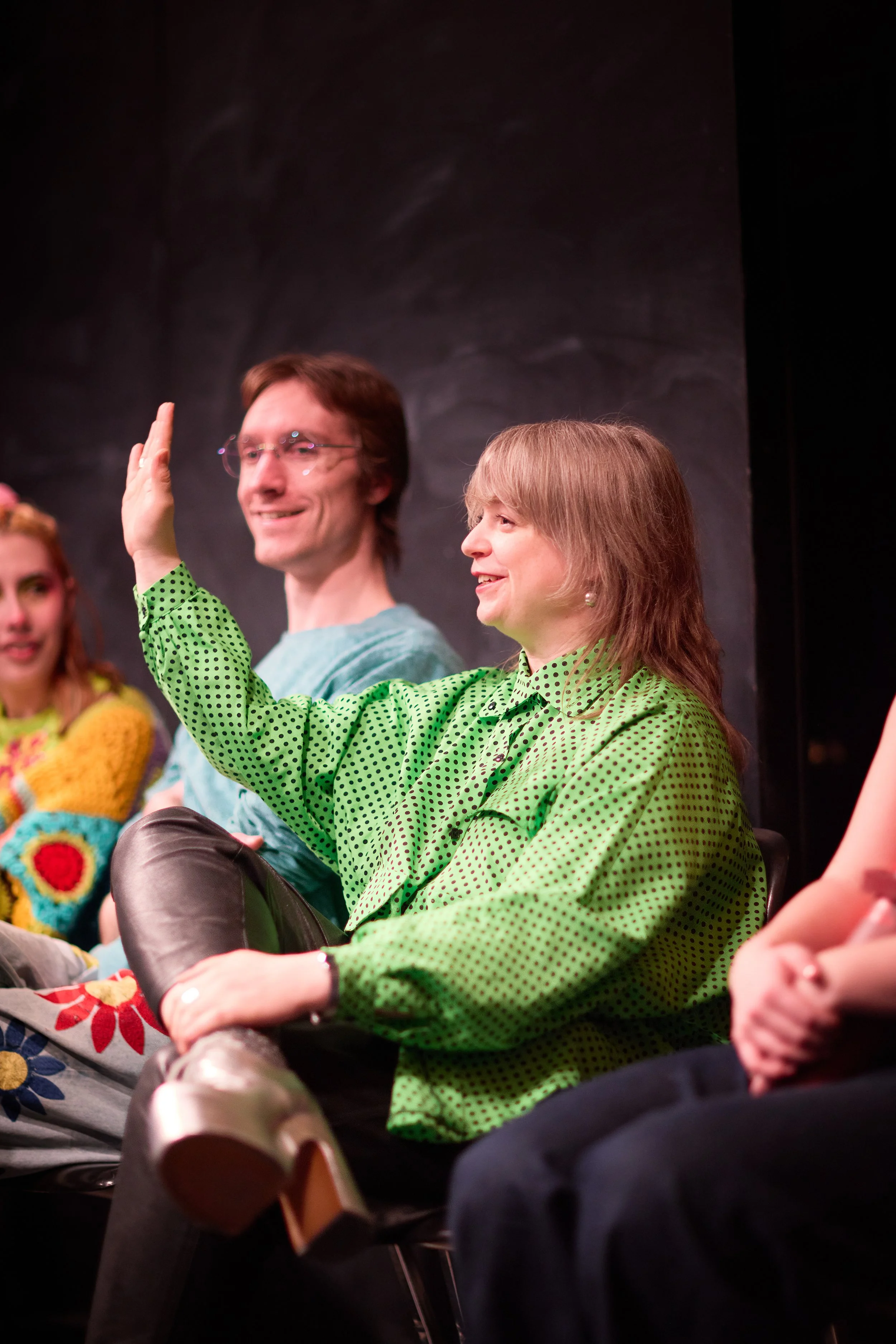 Groupe de personnes assises lors d'une réunion ou d'un événement, dont une femme en pull vert à pois qui lève la main, dans un environnement intérieur avec fond noir.