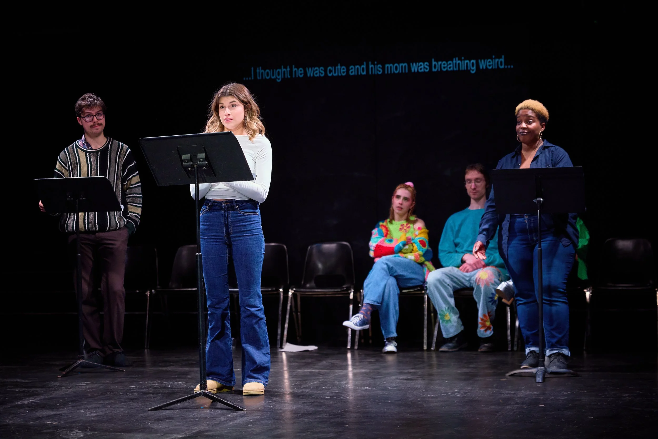 Un groupe de jeunes acteurs sur scène, certains assis et d'autres en train de réciter leur texte avec des supports. Il y a une citation en anglais affichée en haut, disant : « ...je pensais qu'il était mignon et que sa maman respirait bizarre... »