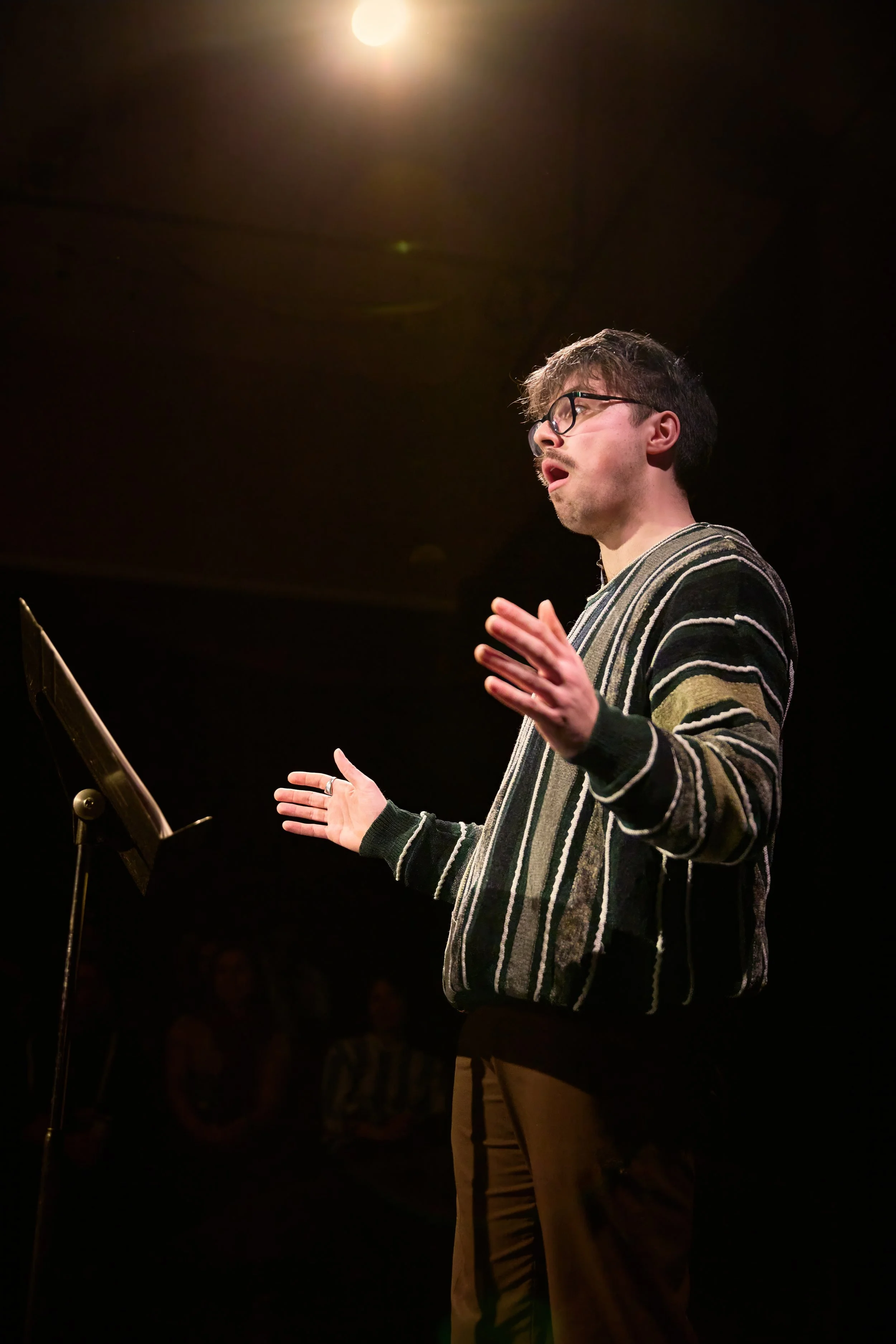 Un jeune homme avec des lunettes, chantant ou parlant, debout devant un pupitre avec un fond sombre et un éclairage au plafond.